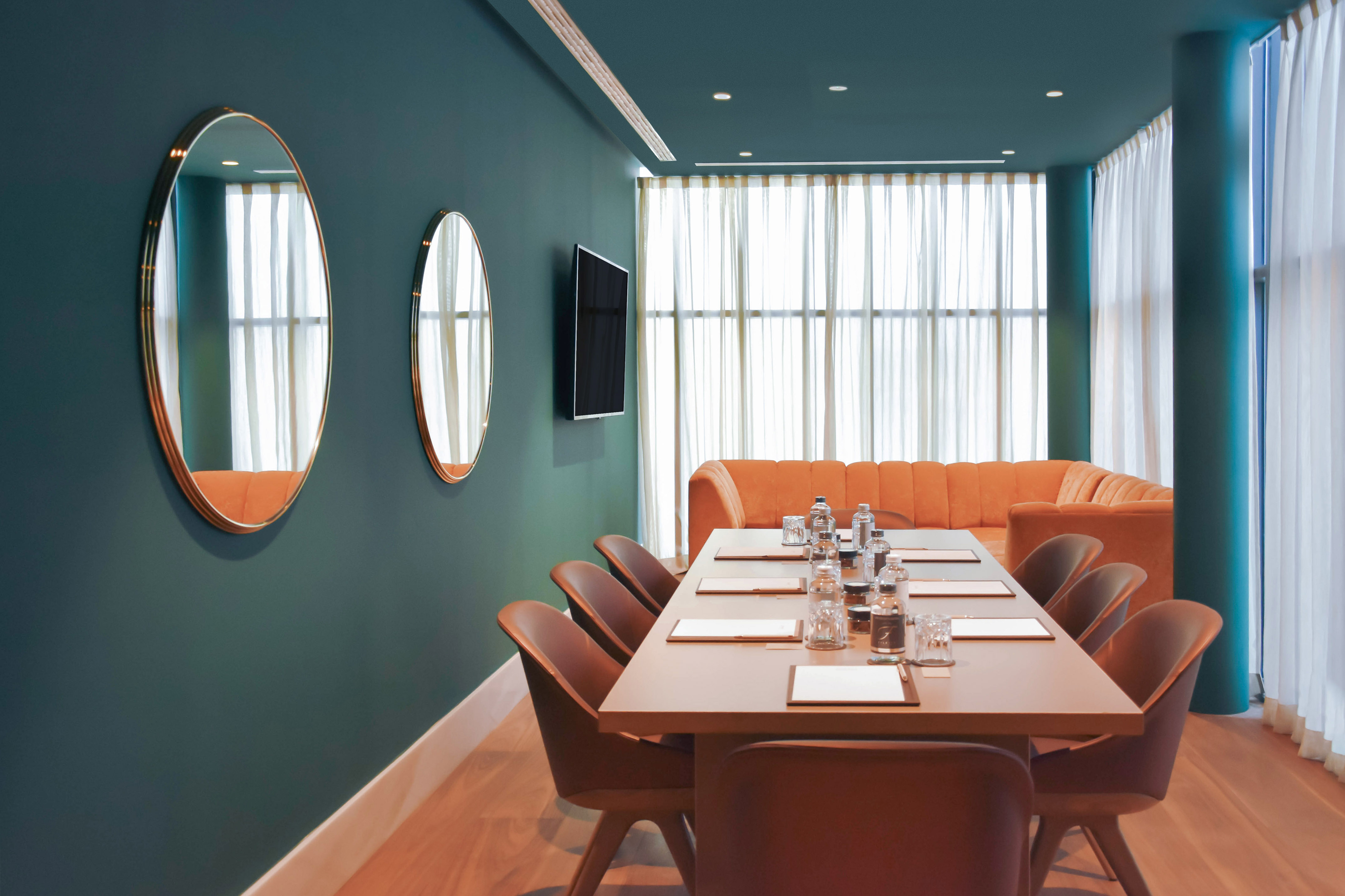 Modern meeting room with teal walls, long table, orange sofa, and large windows.