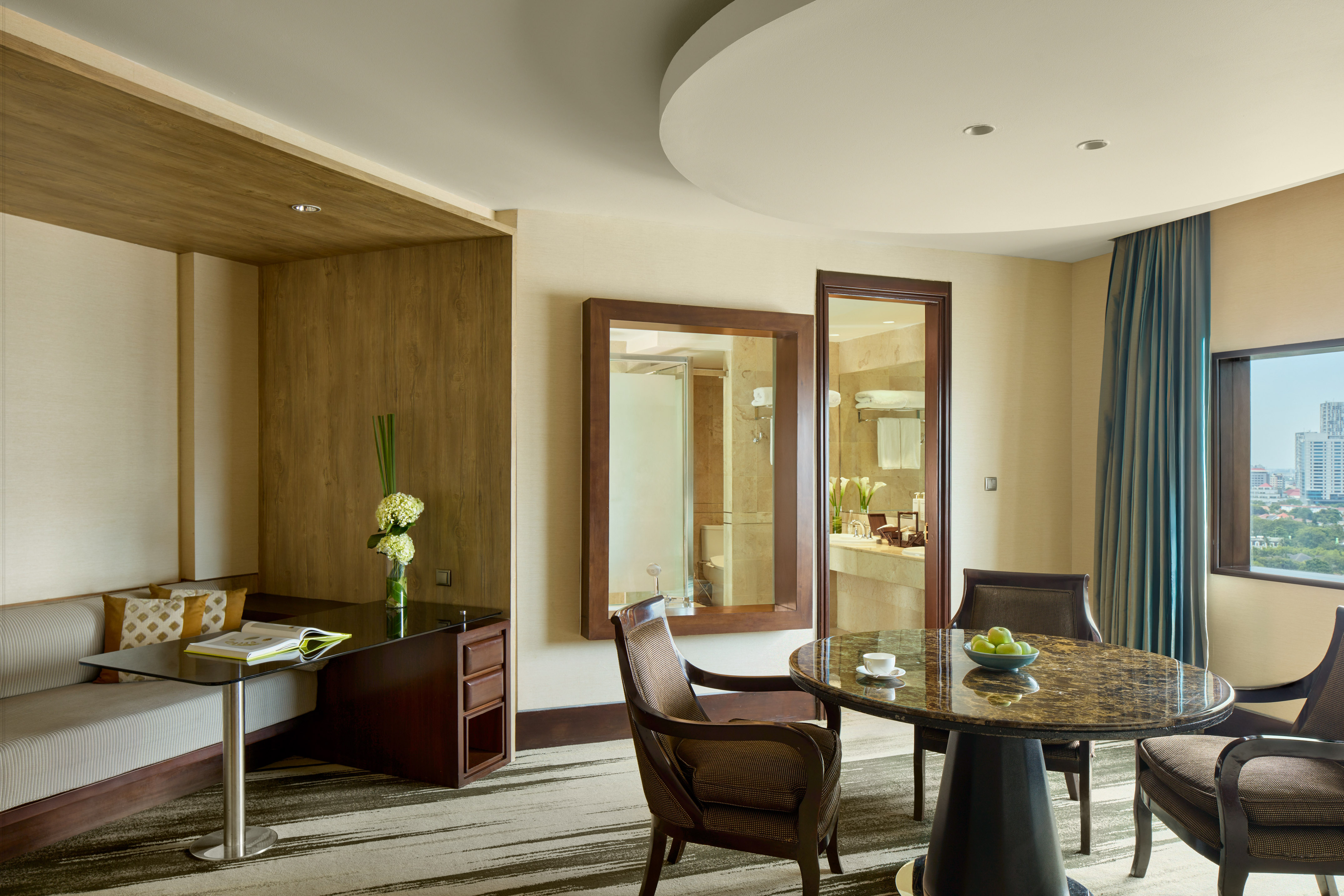 Modern hotel suite living area with desk, dining table, and city view.