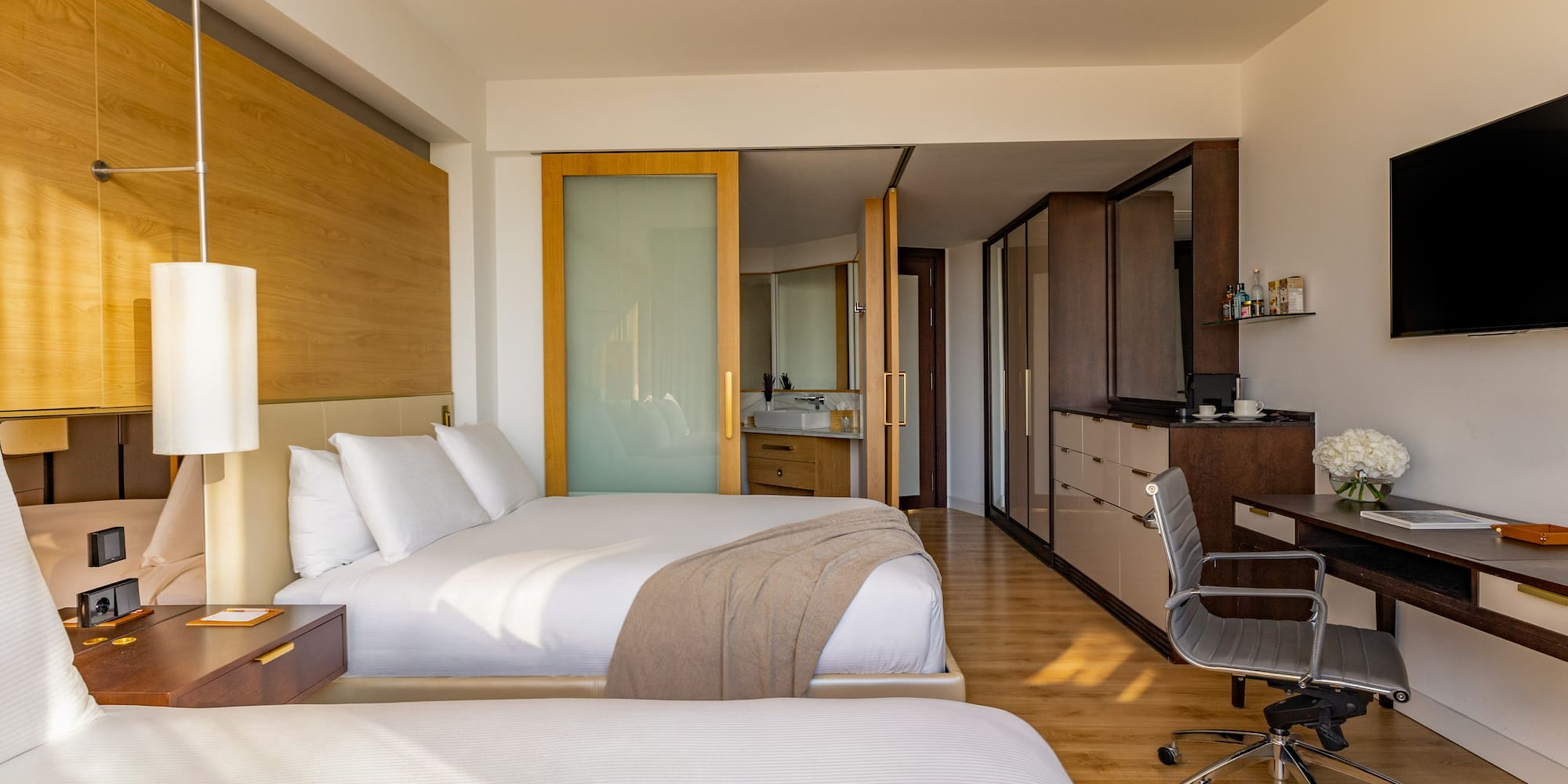Modern hotel room with two beds, workstation, and open-plan bathroom.