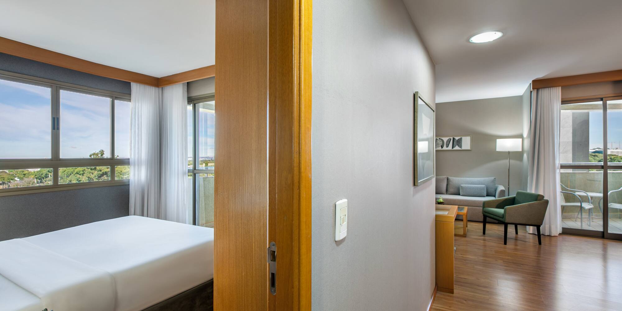 Bright hotel suite featuring bedroom, living room, large windows, and balcony.