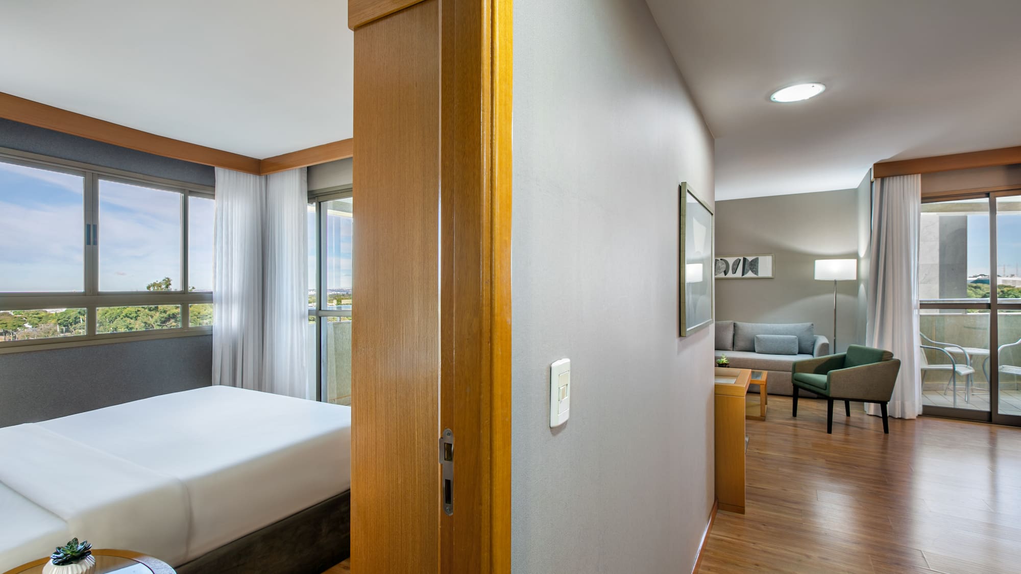 Bright hotel suite featuring bedroom, living room, large windows, and balcony.