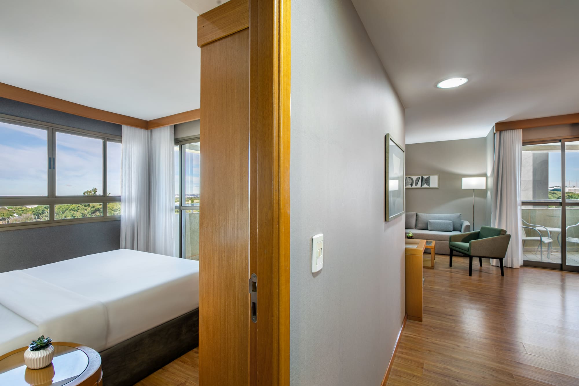 Bright hotel suite featuring bedroom, living room, large windows, and balcony.