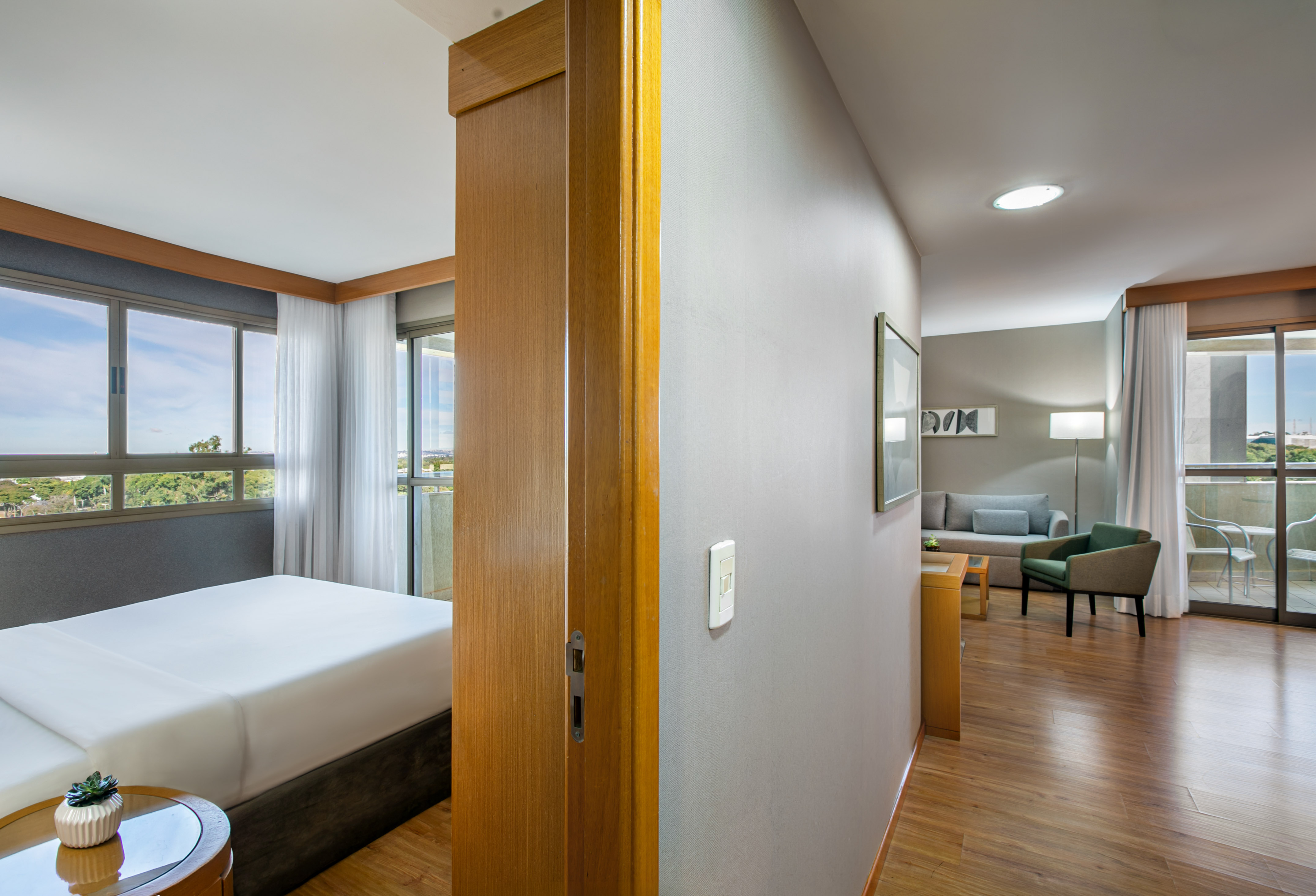 Bright hotel suite featuring bedroom, living room, large windows, and balcony.