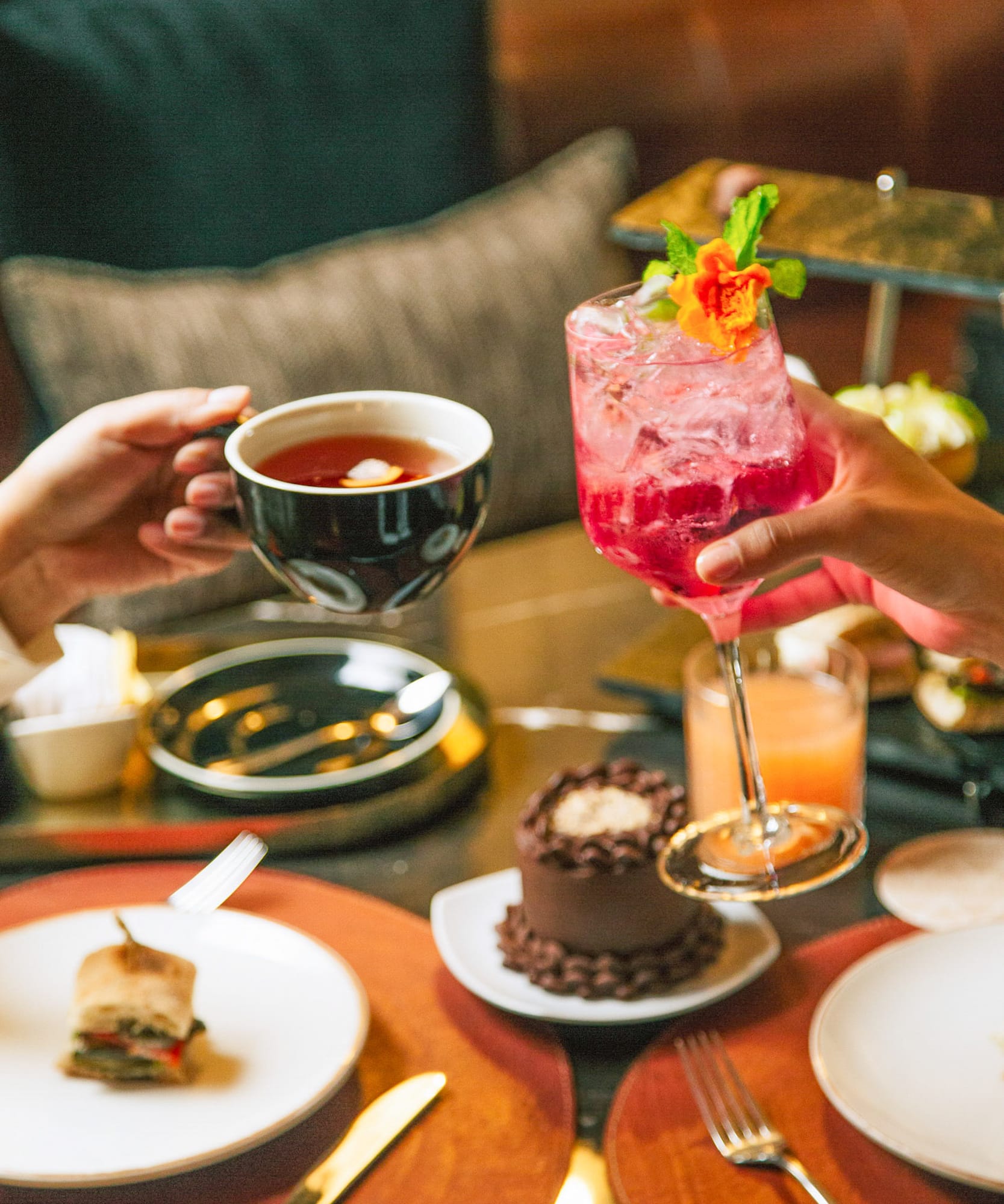 Manos brindando con té y cóctel rosa en una elegante merienda.