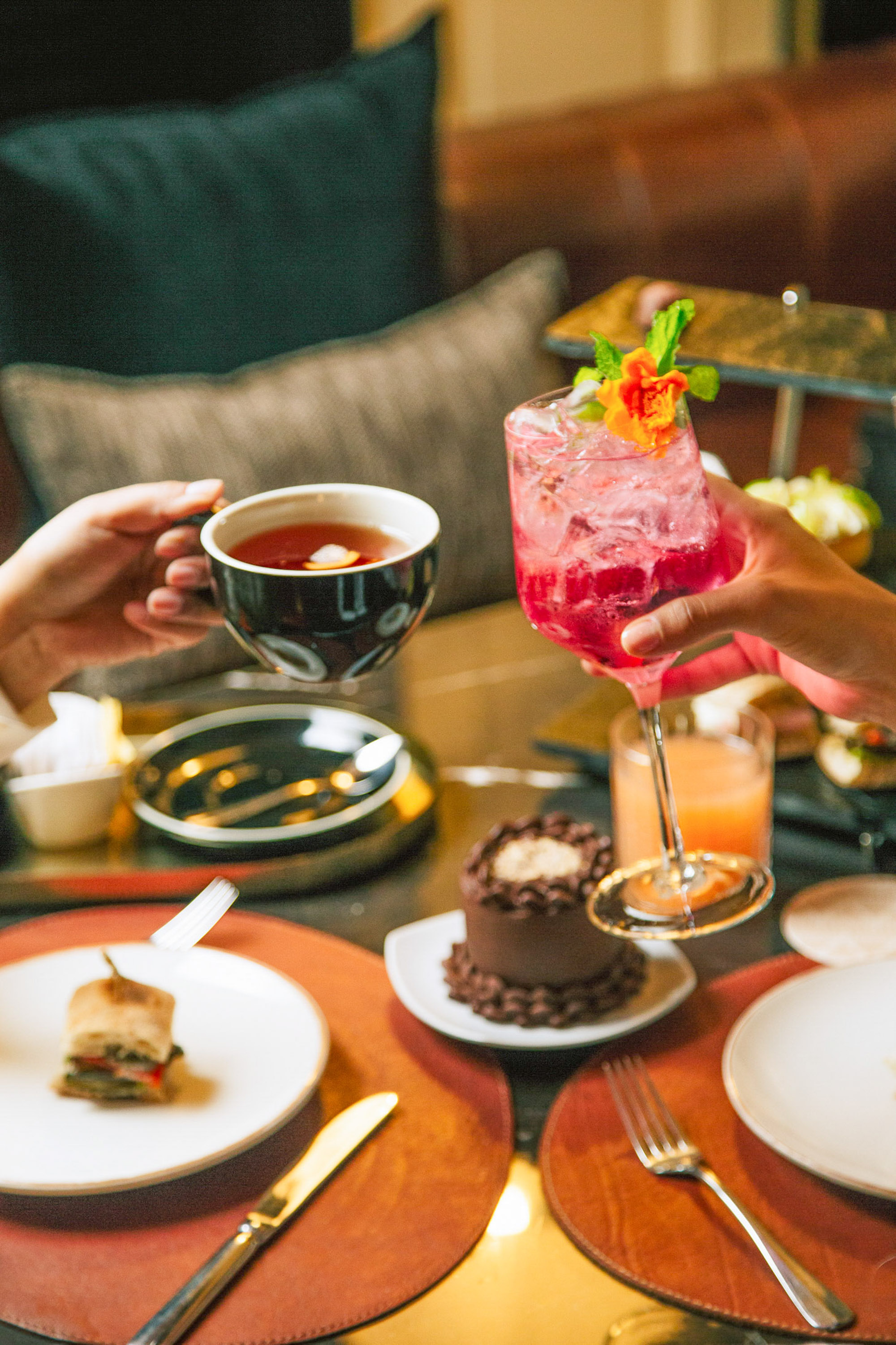 Manos brindando con té y cóctel rosa en una elegante merienda.