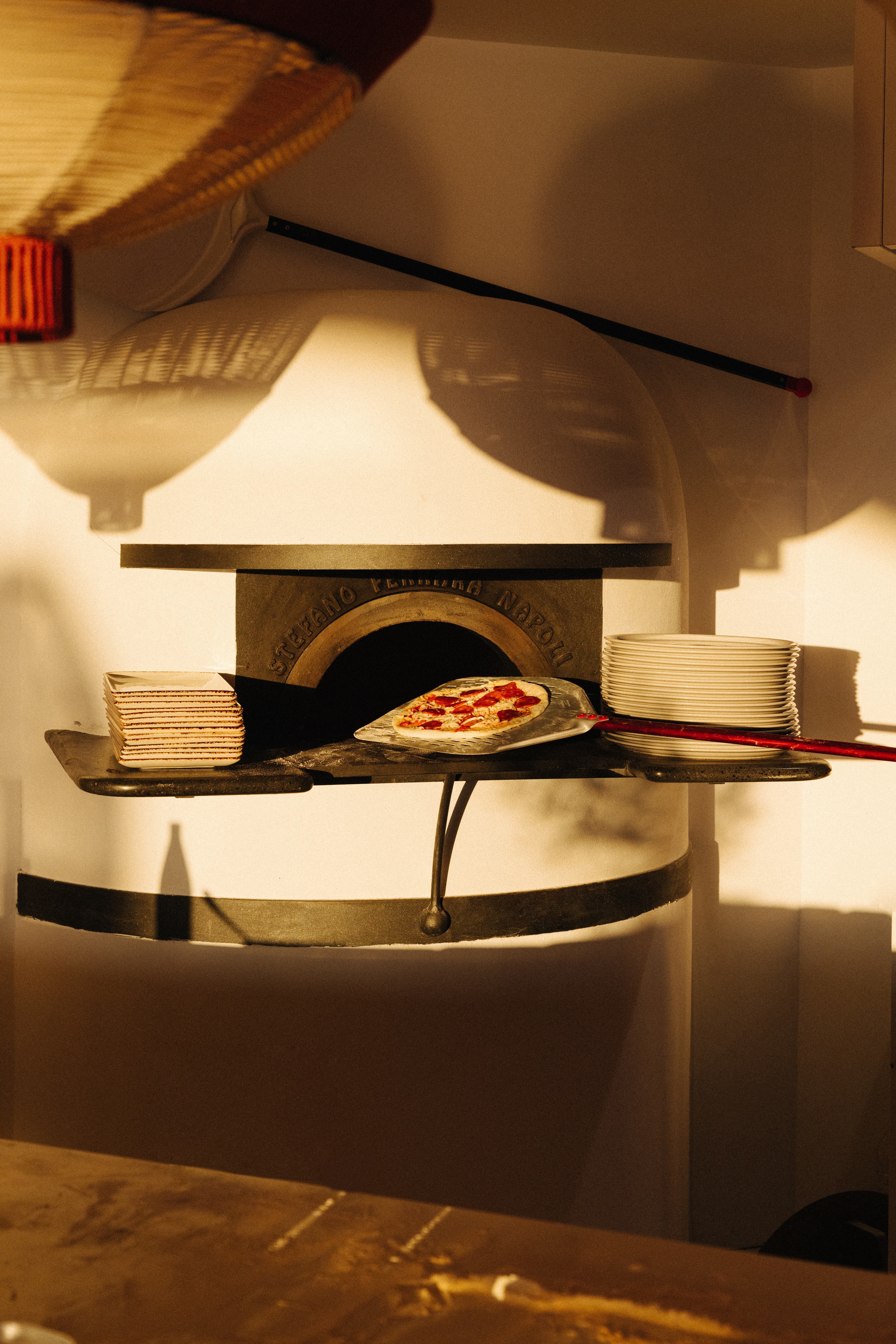 Uncooked pepperoni pizza on peel near Napoli pizza oven, stacked plates.