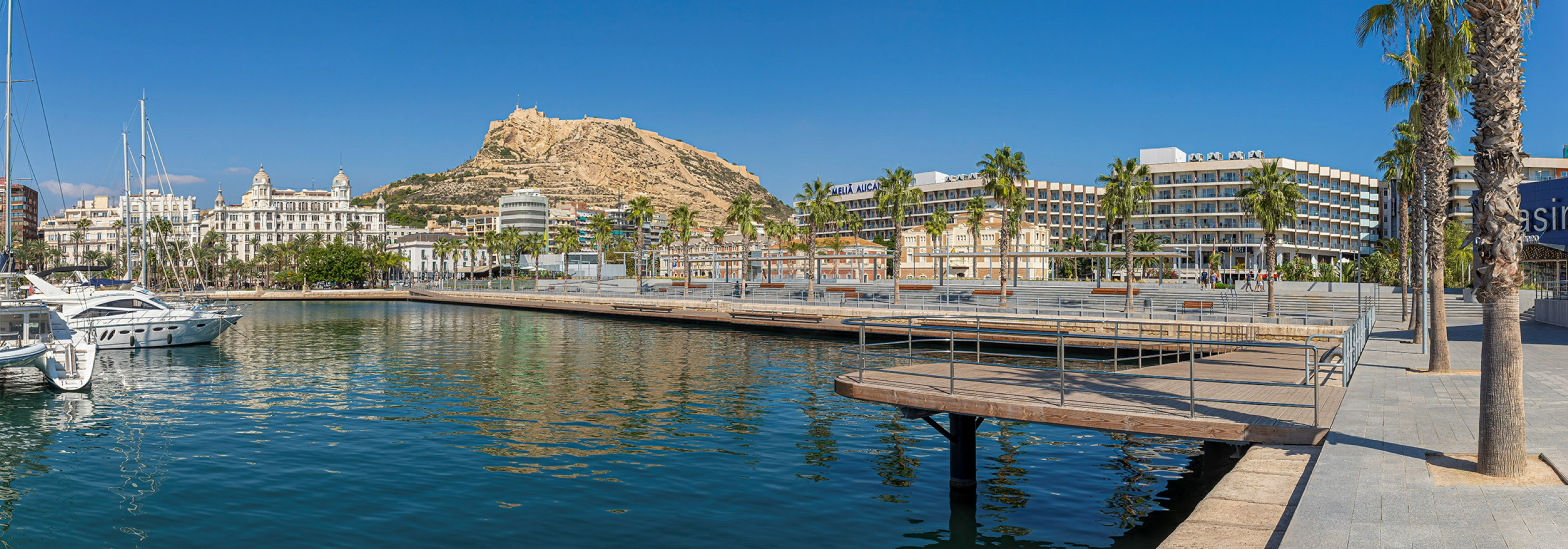 Alicante marina with yachts, city buildings, palm trees, and Santa Barbara Castle.