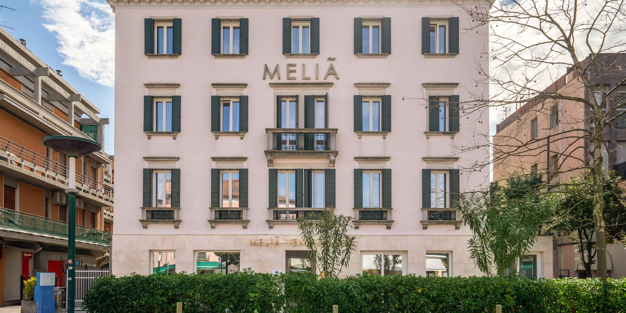 Melia hotel building with light facade, green shutters, balcony, and street-level hedge.