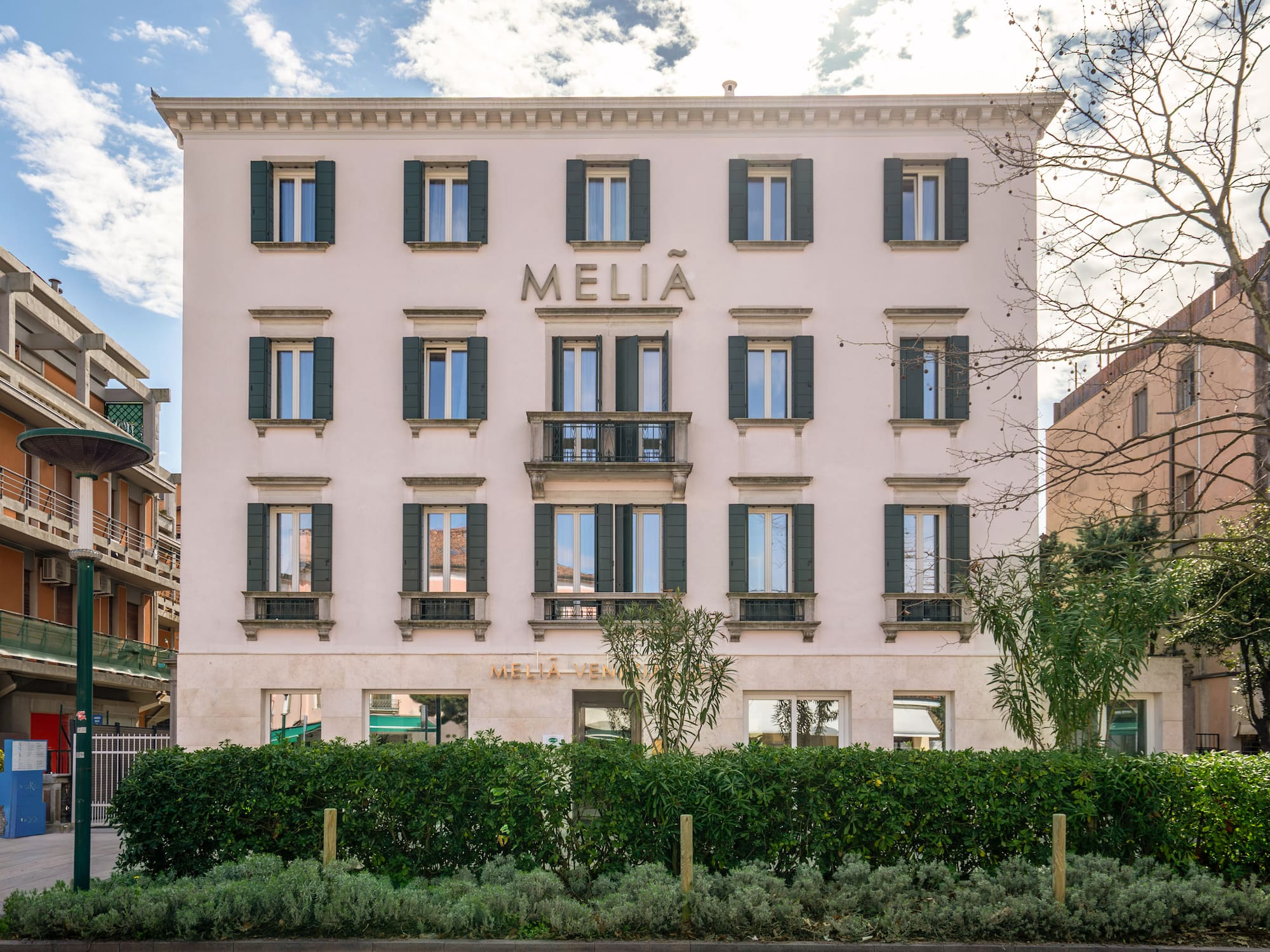 Melia hotel building with light facade, green shutters, balcony, and street-level hedge.
