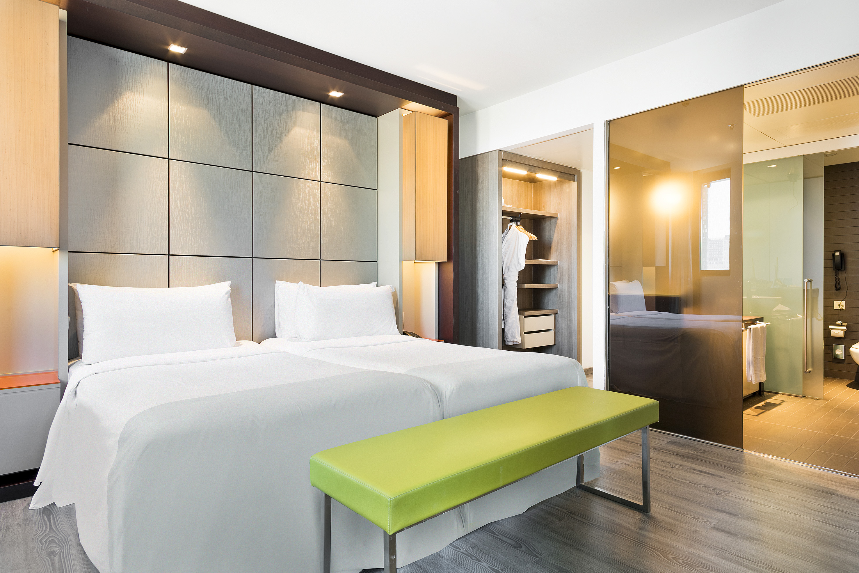 Modern hotel room with twin beds, green bench, and glass-walled bathroom.