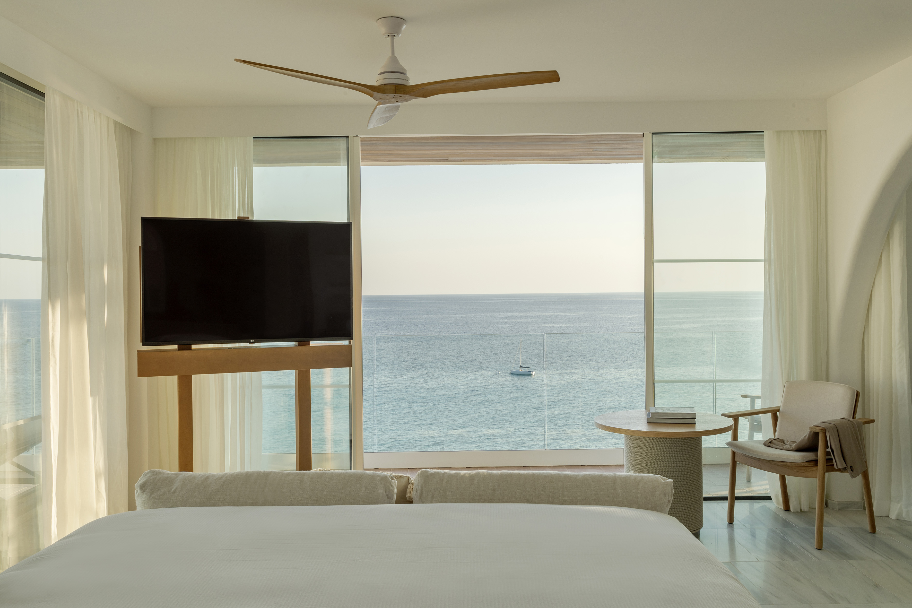 Bright ocean view bedroom suite with bed, TV, and distant sailboat.