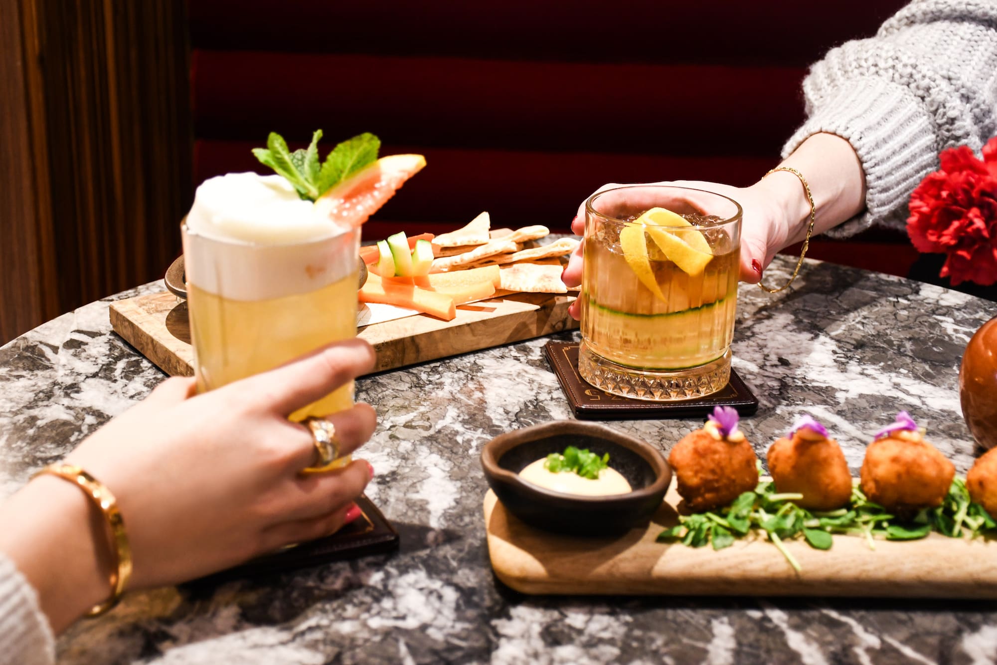 Two hands holding craft cocktails and appetizers on a marble table at a bar.