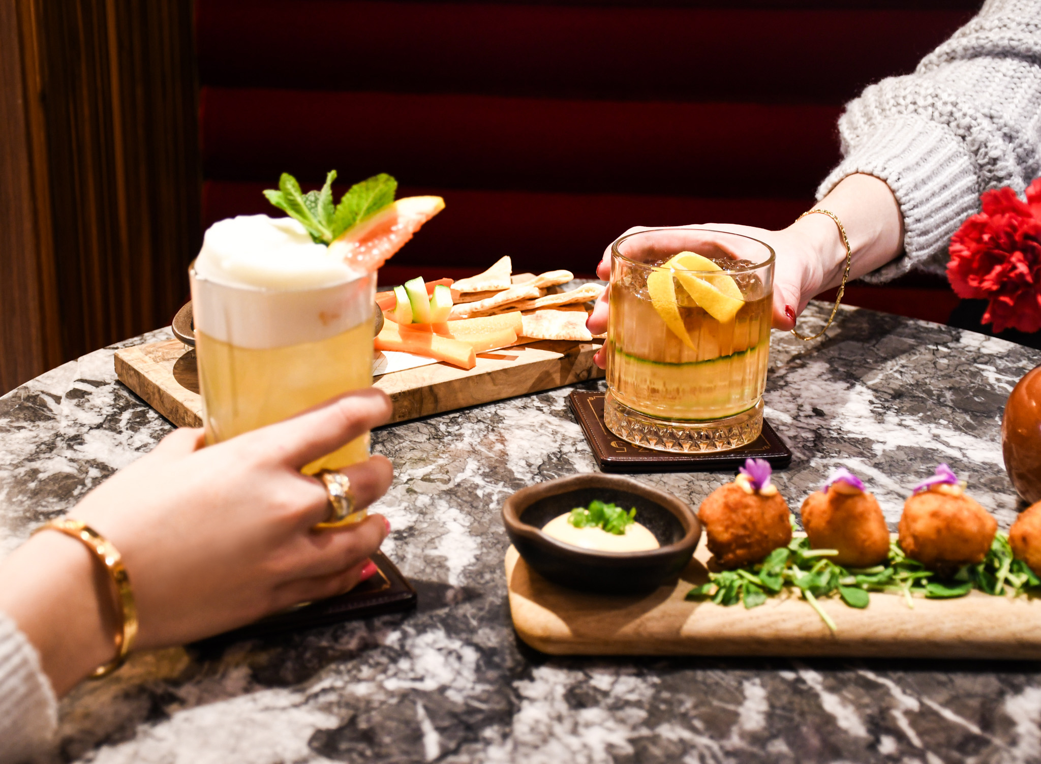 Two hands holding craft cocktails and appetizers on a marble table at a bar.