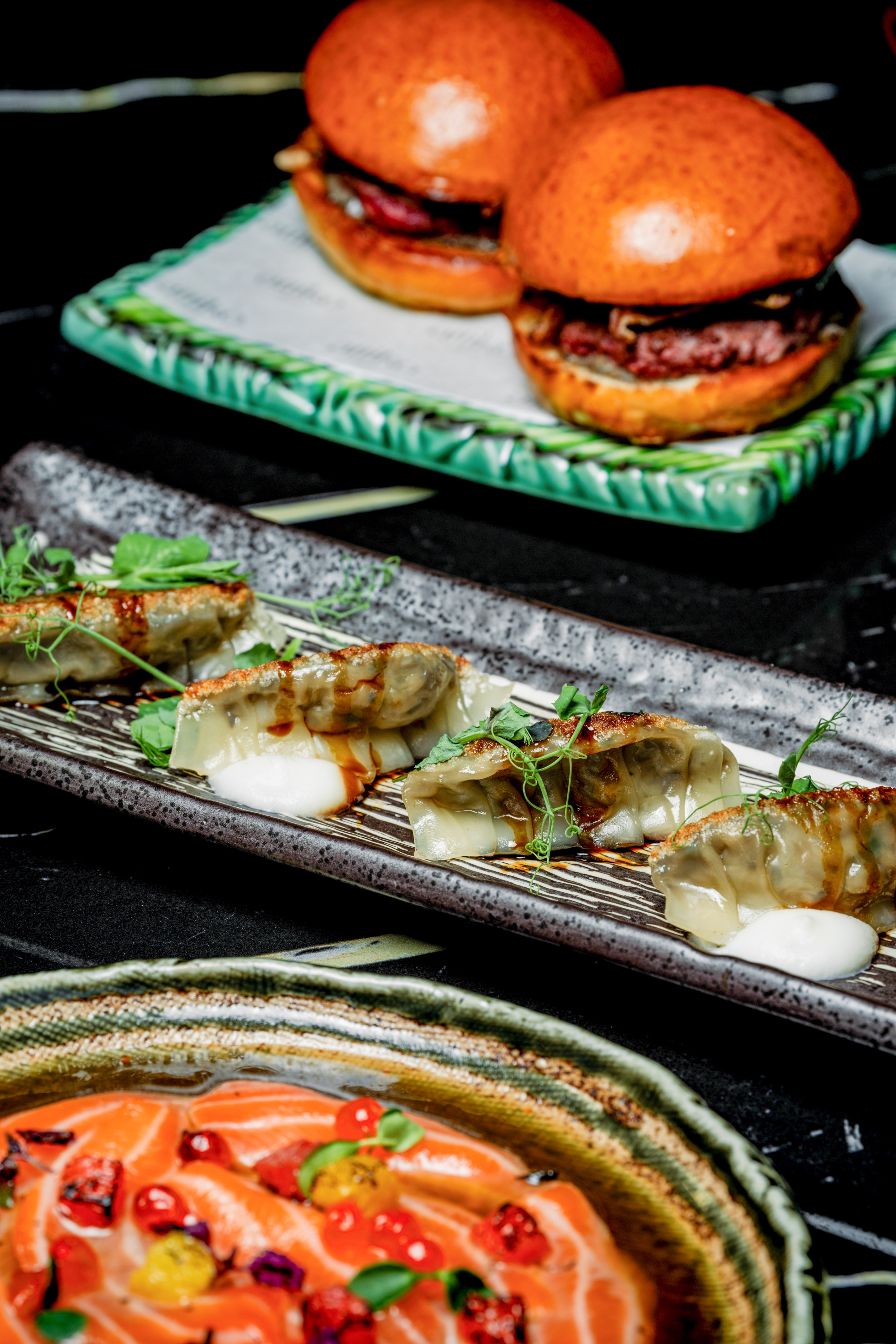 Pan-fried gyoza, fresh salmon, and mini burgers on dark plates.