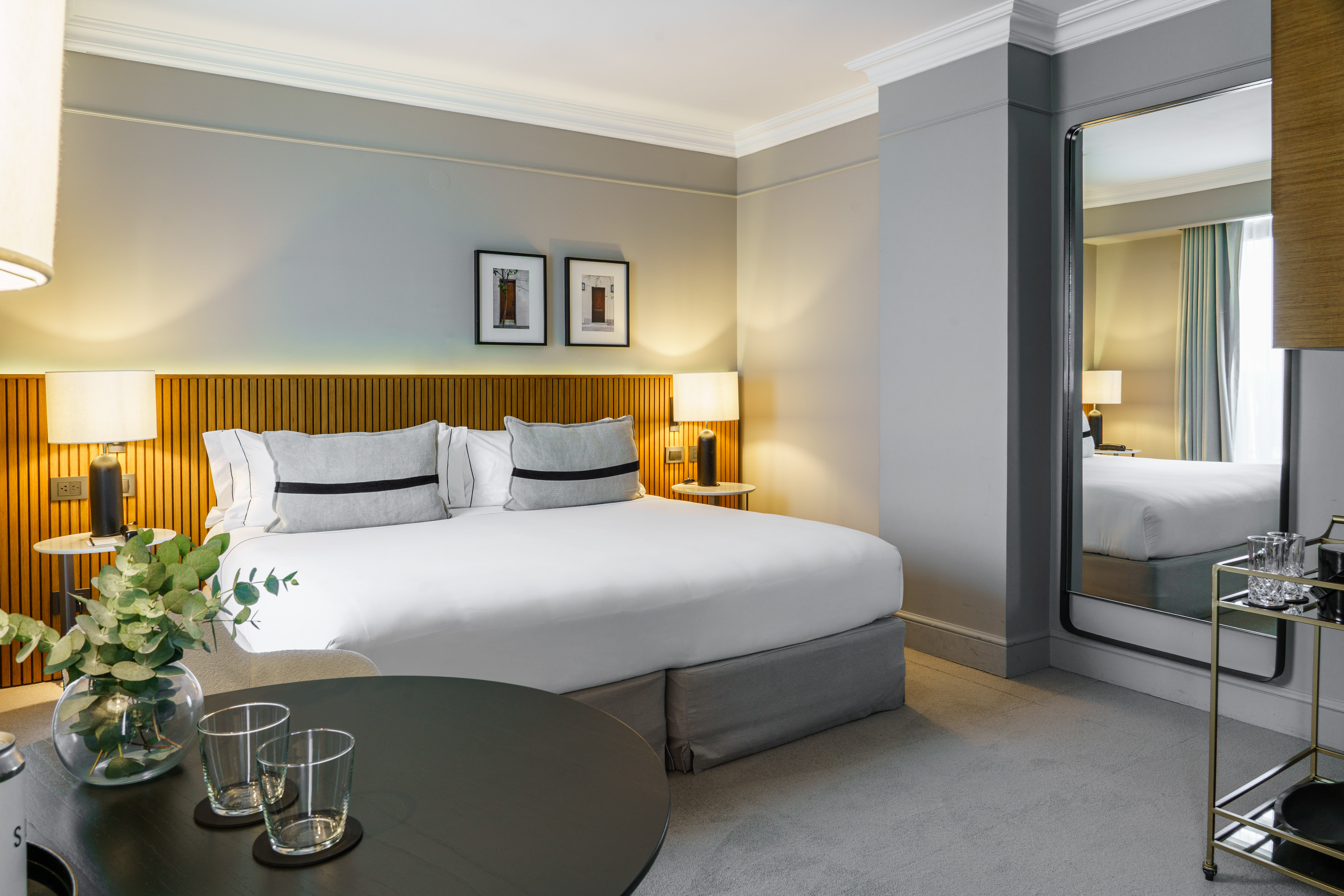 Modern hotel bedroom with king bed, wooden headboard, elegant decor, and mirror.