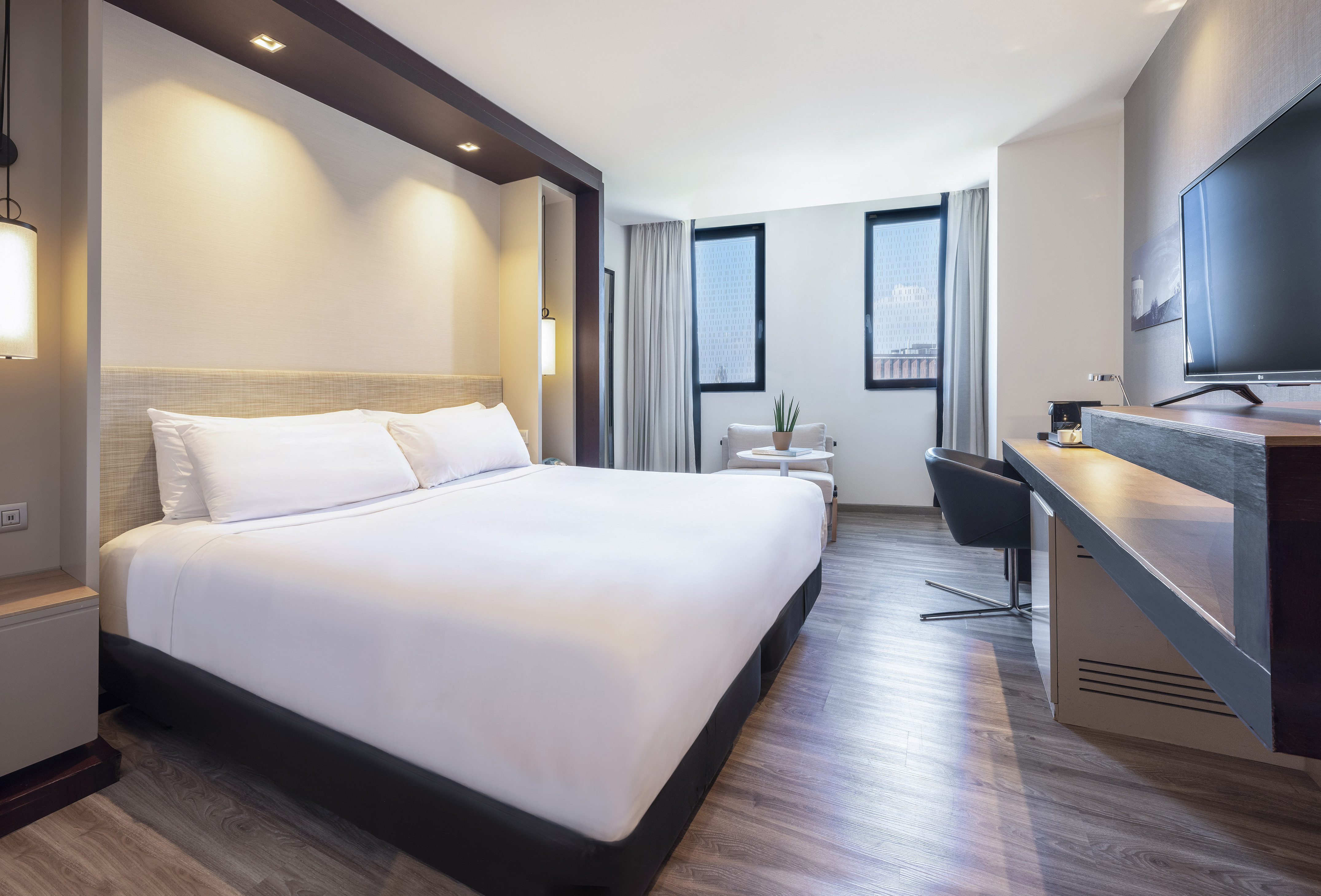 Modern hotel room with large bed, desk, TV, and bright windows.
