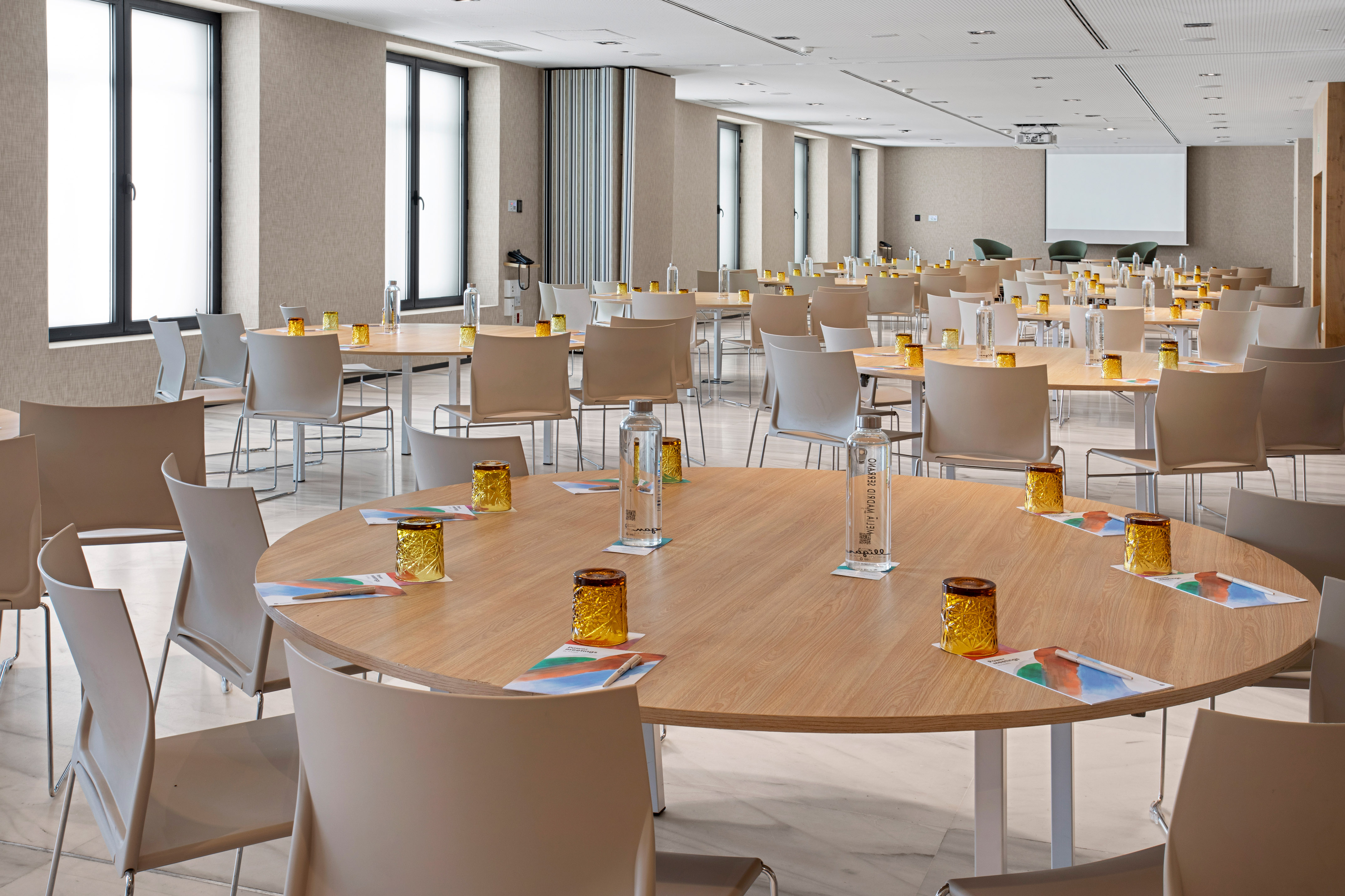Modern meeting room with round tables, chairs, water, and projector screen.