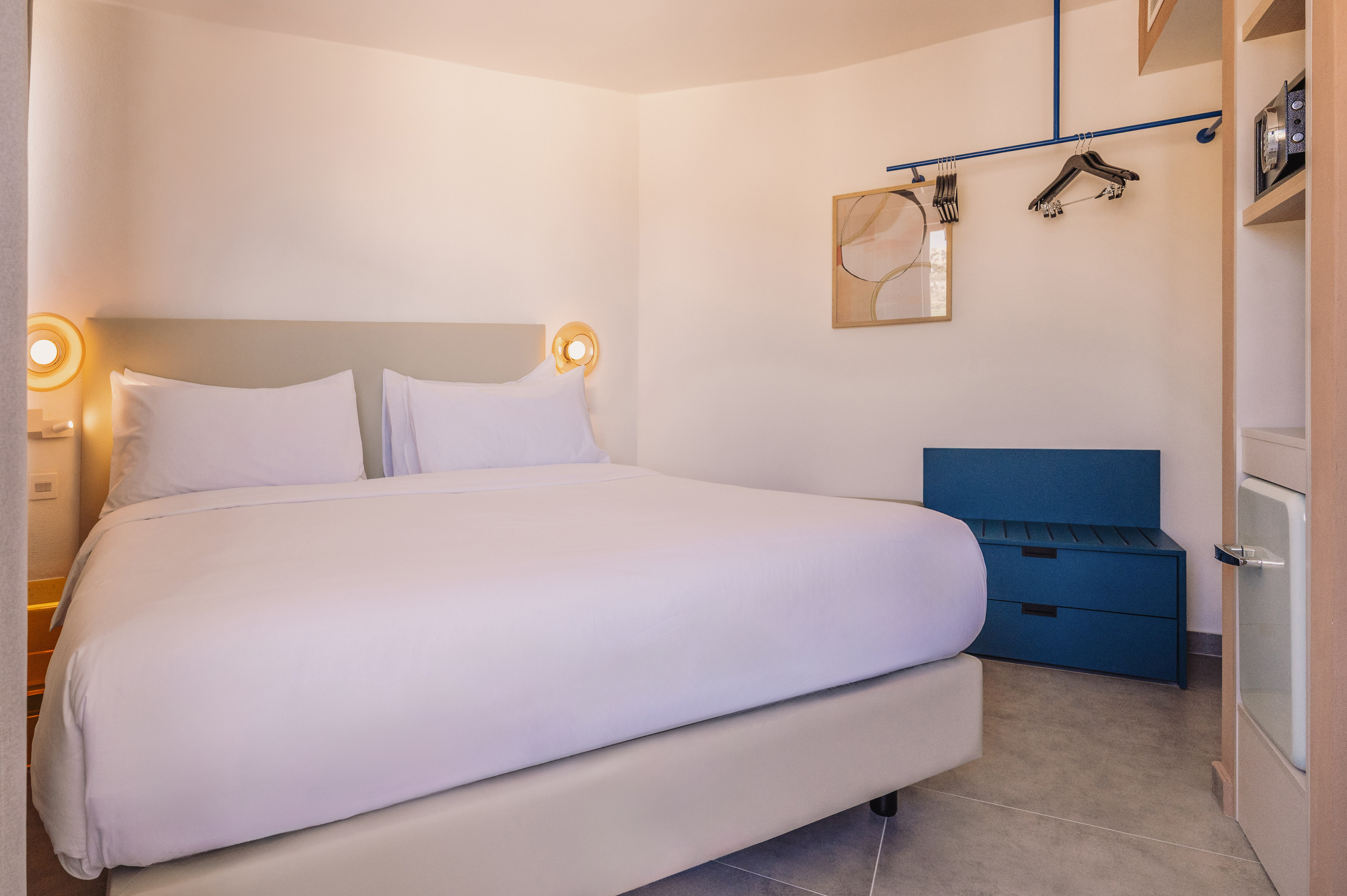 Modern hotel room with white bed, blue storage bench, and clothes rack.