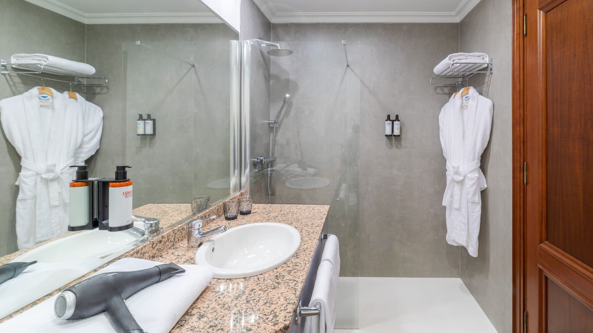 Modern hotel bathroom with shower, vanity, mirror, white towels, and bathrobes.