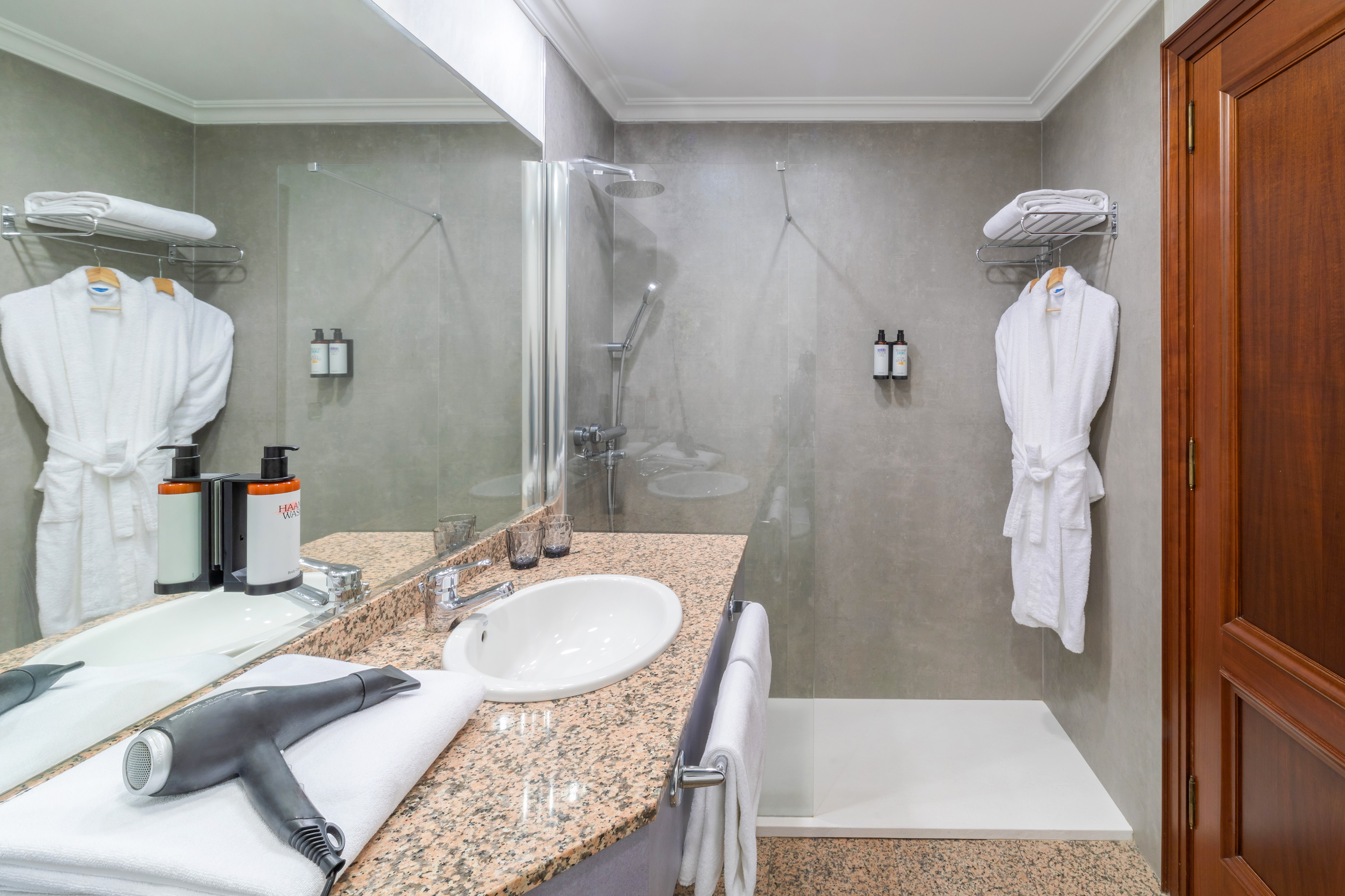 Modern hotel bathroom with shower, vanity, mirror, white towels, and bathrobes.