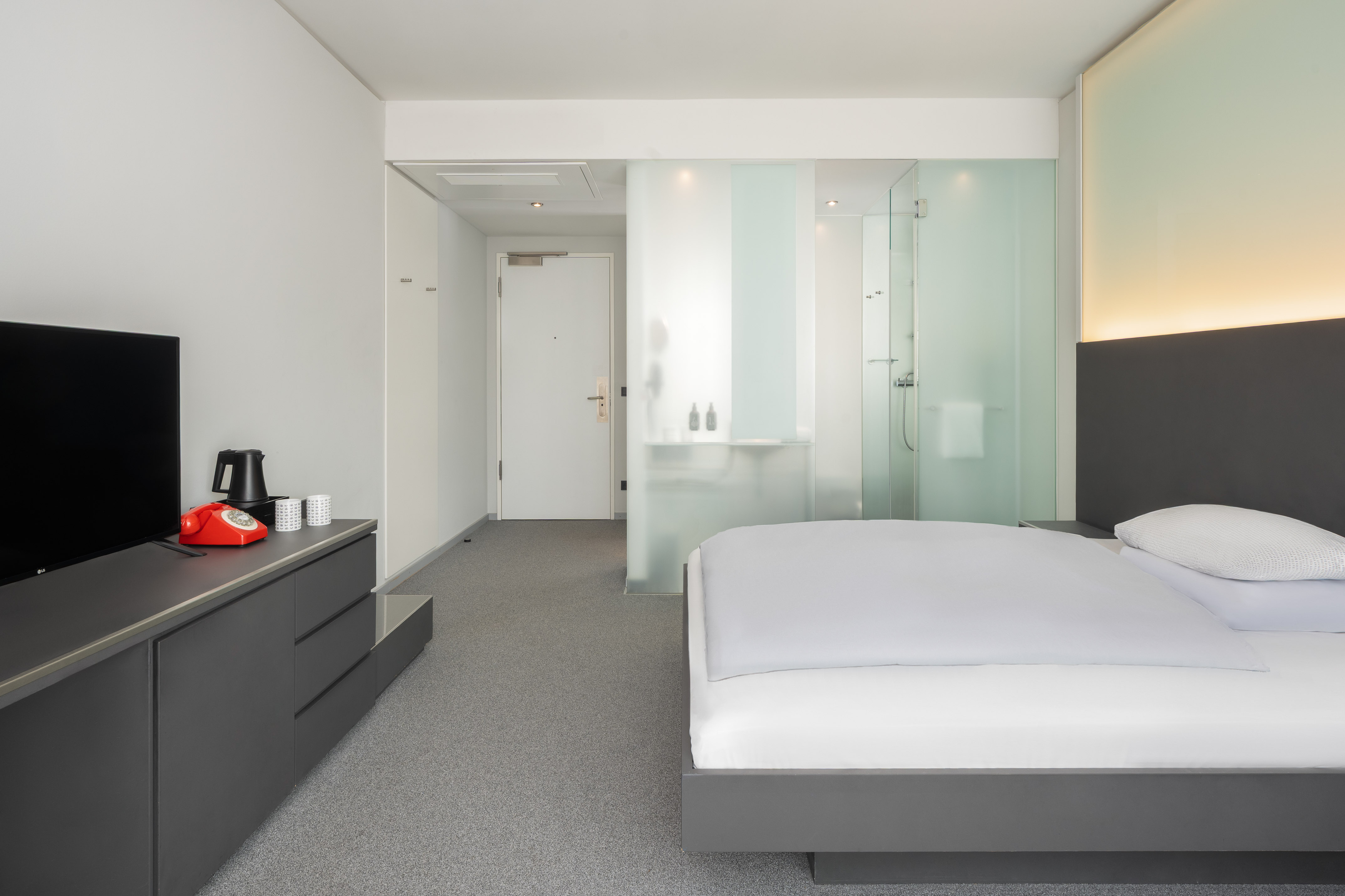 Modern minimalist hotel room with bed, glass shower, TV, and red telephone.