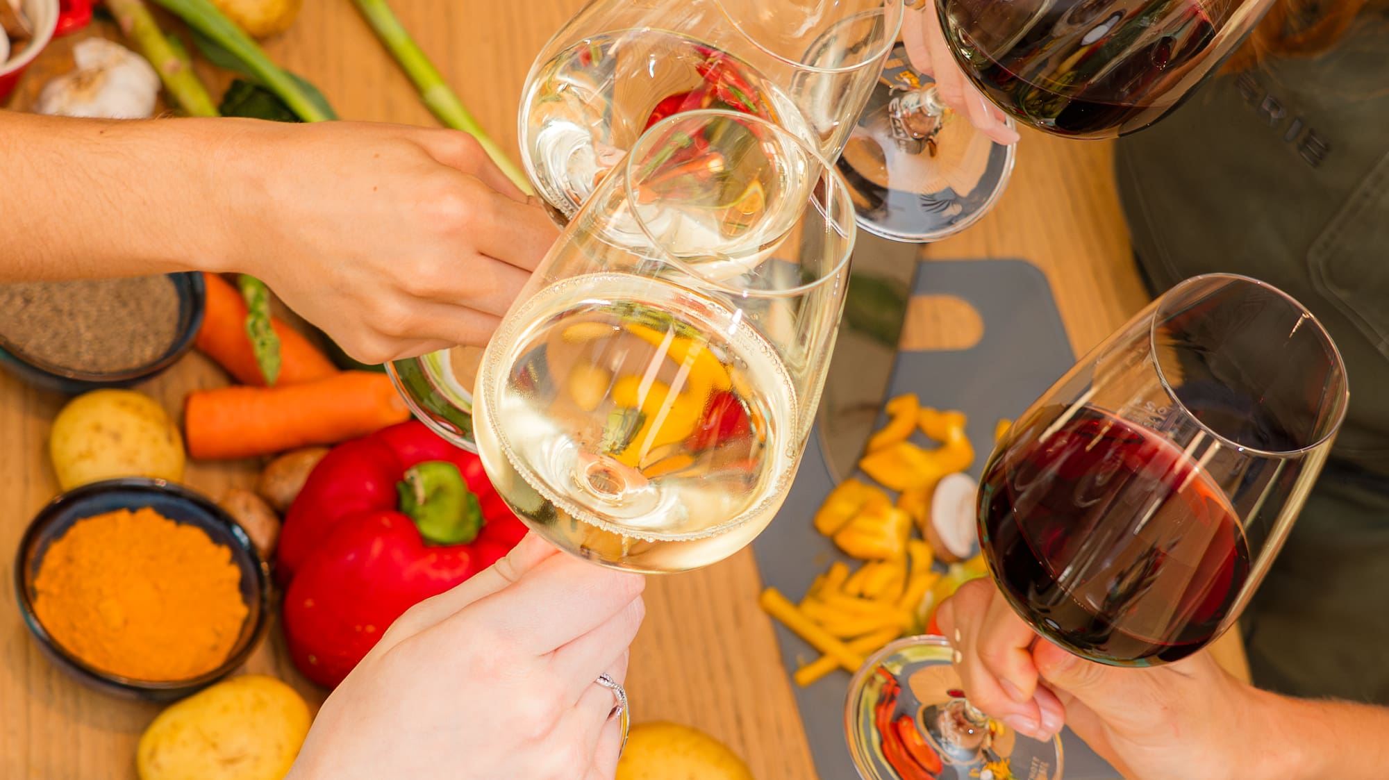 Four hands clinking wine glasses over fresh kitchen ingredients and spices.