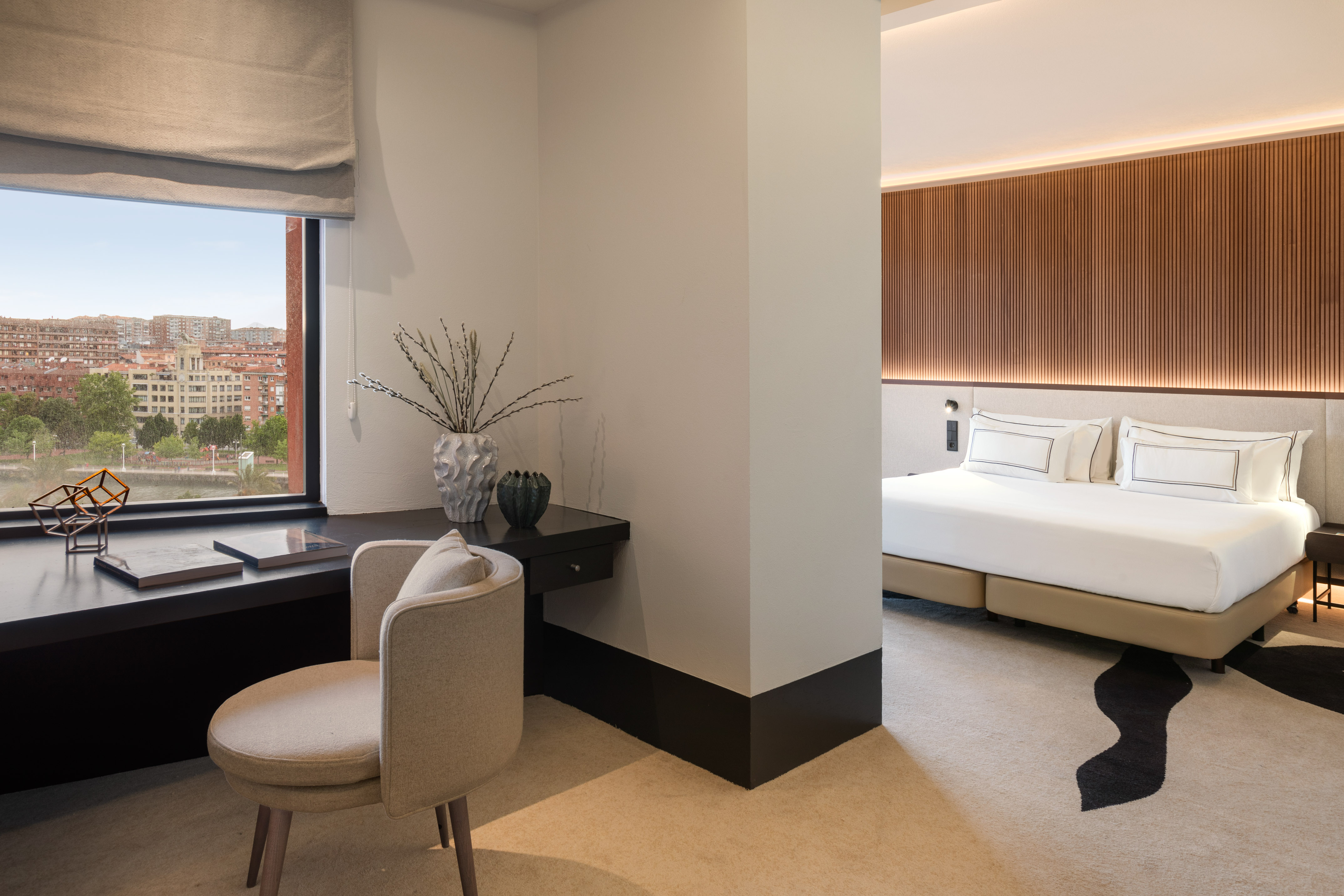 Modern hotel suite with large bed, stylish desk, and city window view.