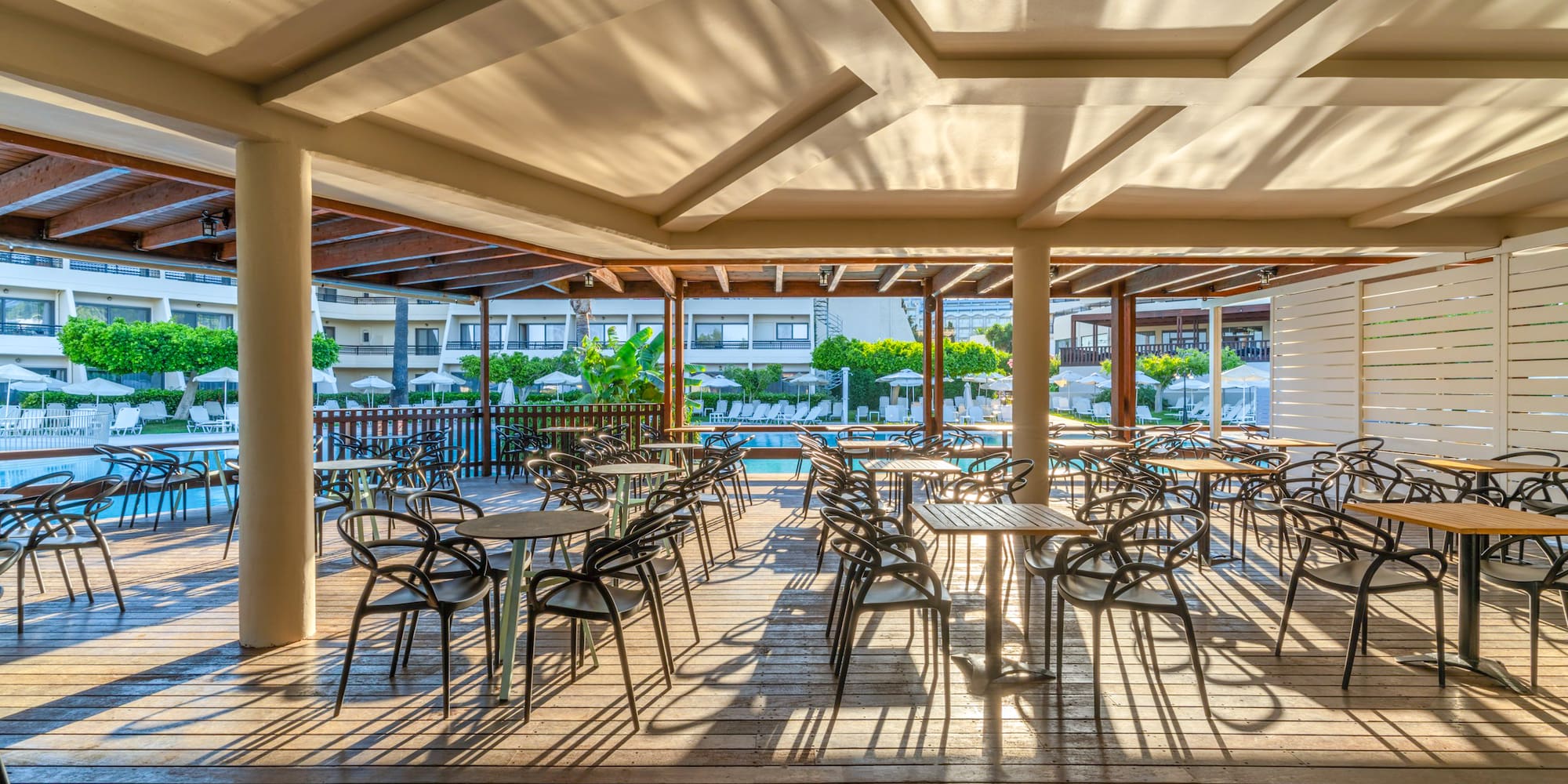 Shaded outdoor restaurant patio with tables, chairs, and hotel pool view.