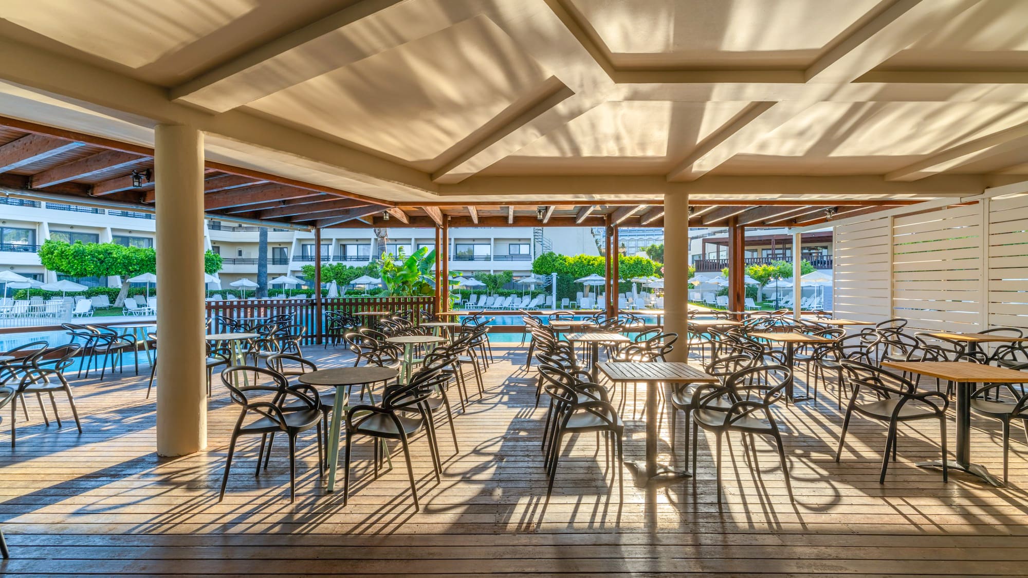 Shaded outdoor restaurant patio with tables, chairs, and hotel pool view.