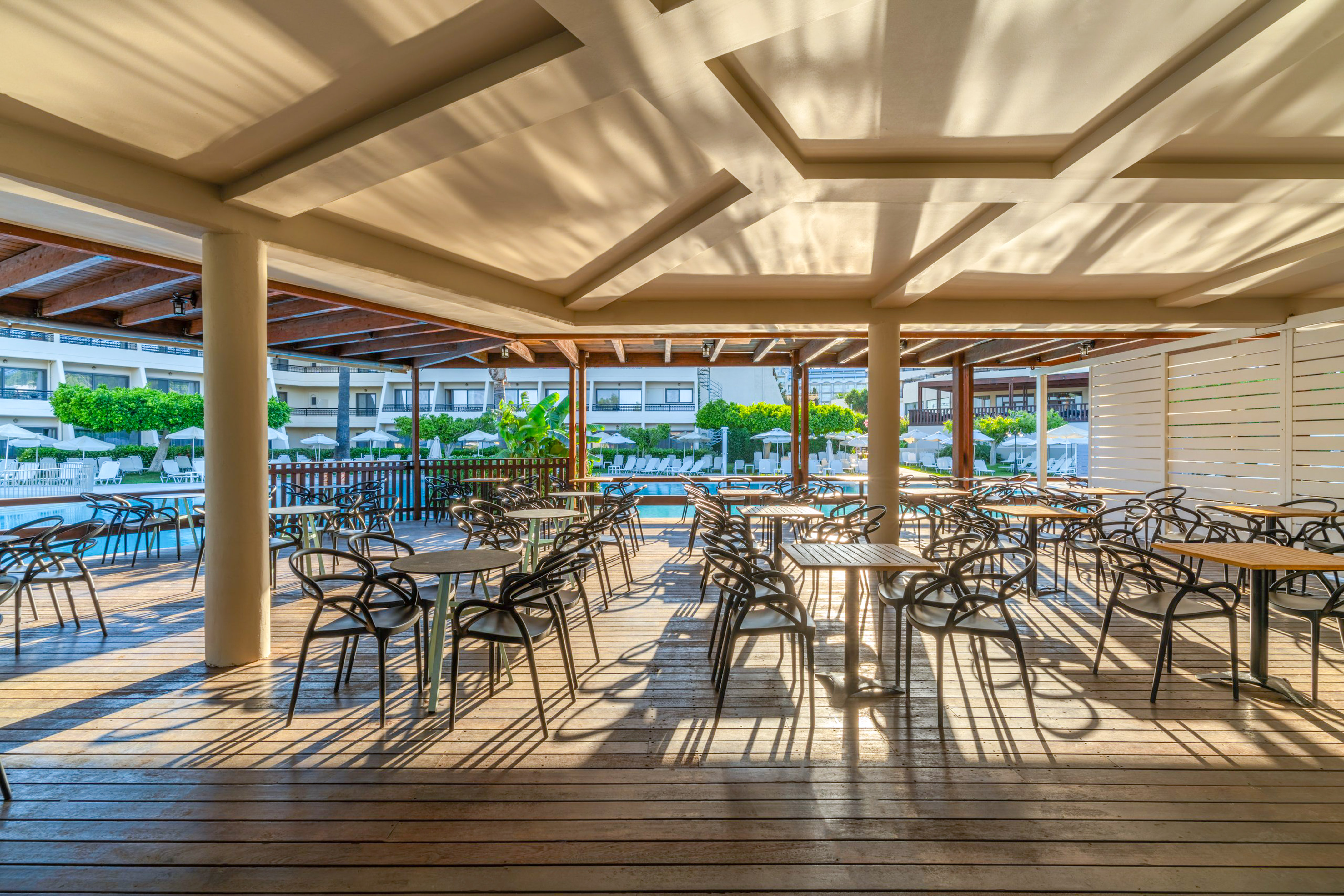 Shaded outdoor restaurant patio with tables, chairs, and hotel pool view.