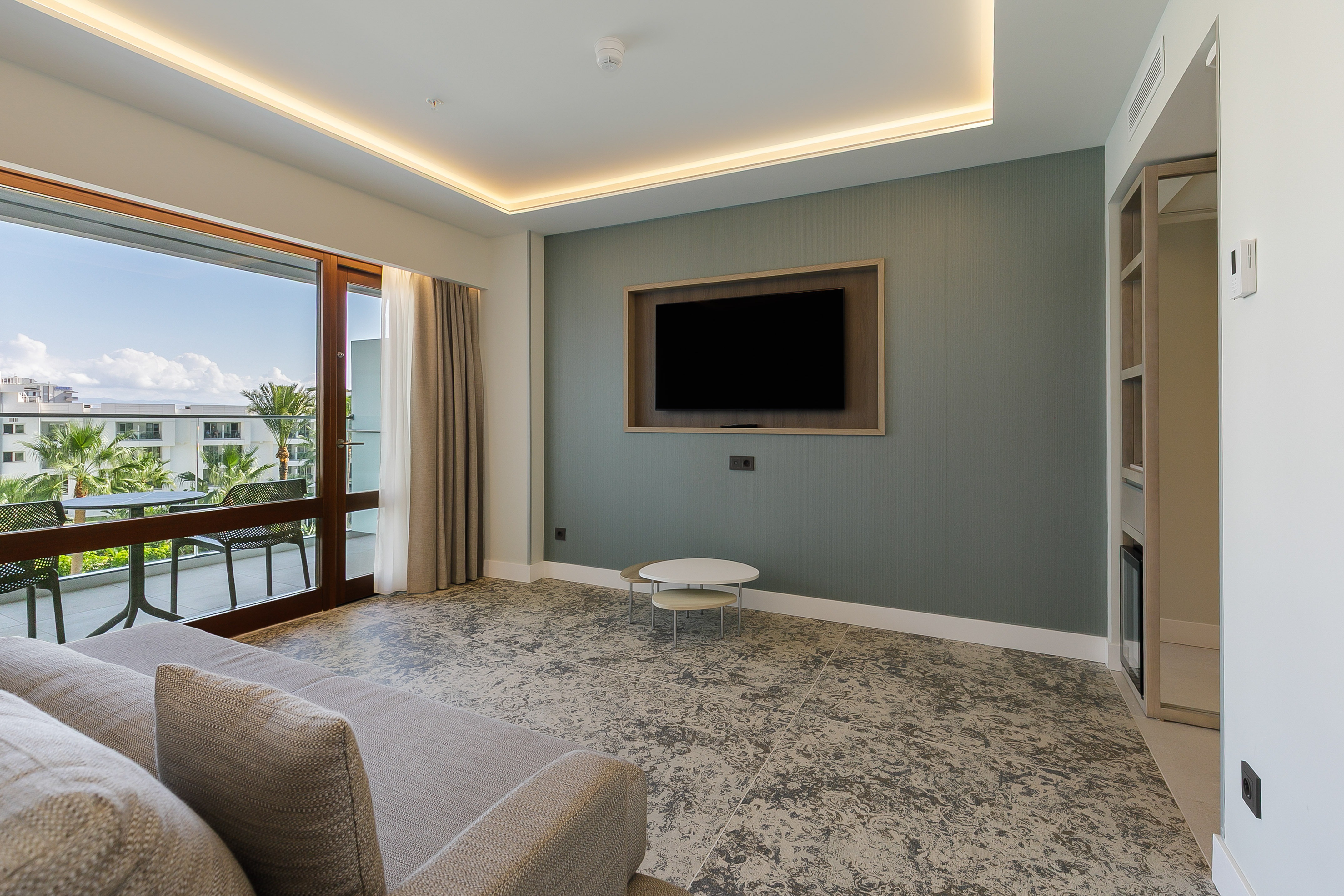 Hotel living room with sofa, wall-mounted TV, and balcony overlooking palm trees.