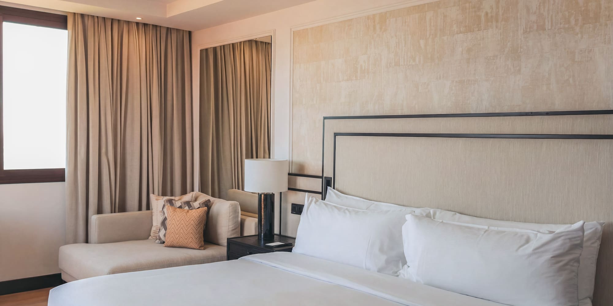 Elegant hotel bedroom with white bed, beige headboard, and chaise lounge by window.