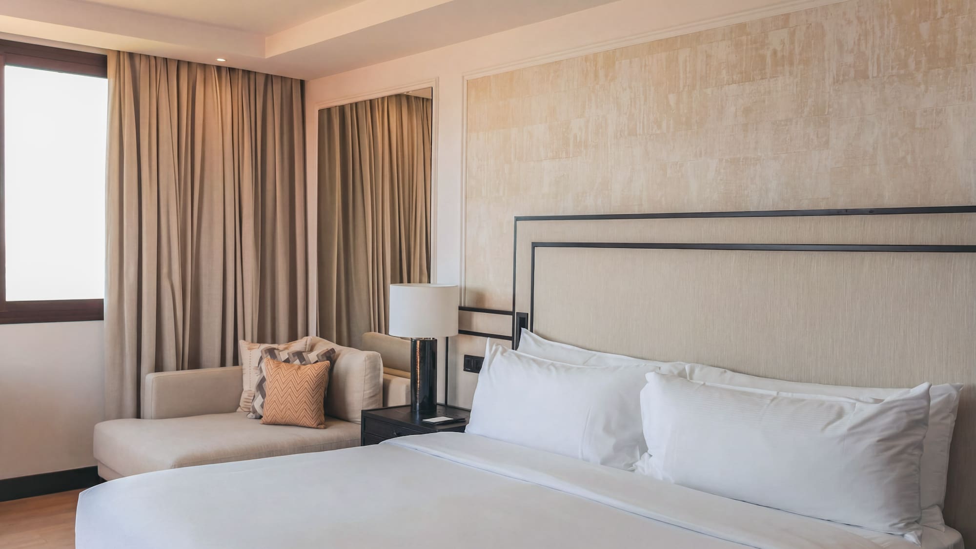 Elegant hotel bedroom with white bed, beige headboard, and chaise lounge by window.