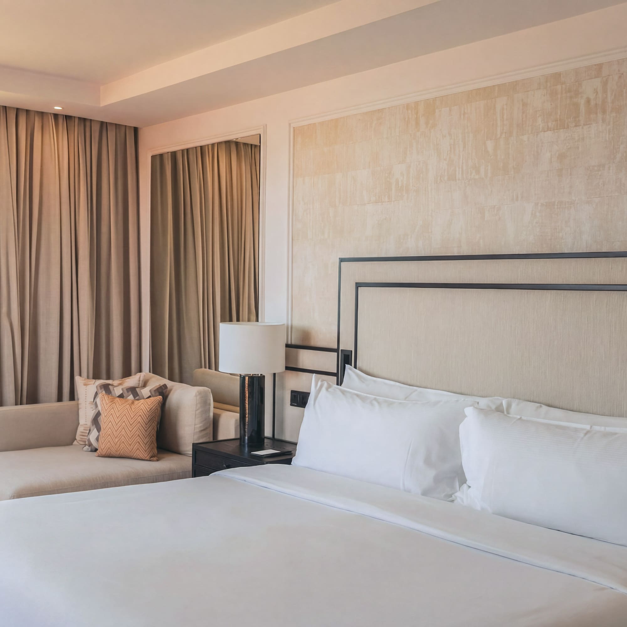 Elegant hotel bedroom with white bed, beige headboard, and chaise lounge by window.