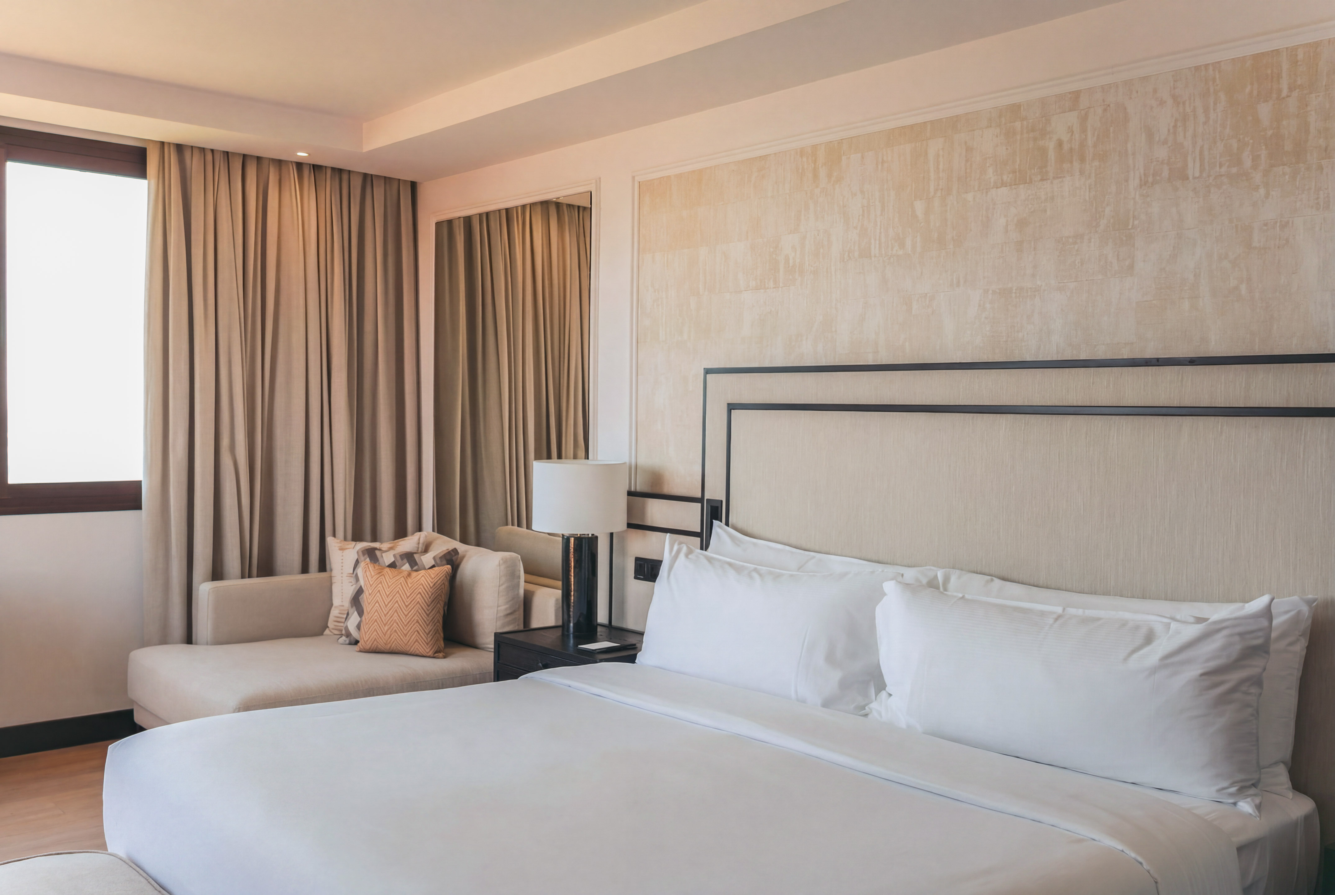 Elegant hotel bedroom with white bed, beige headboard, and chaise lounge by window.