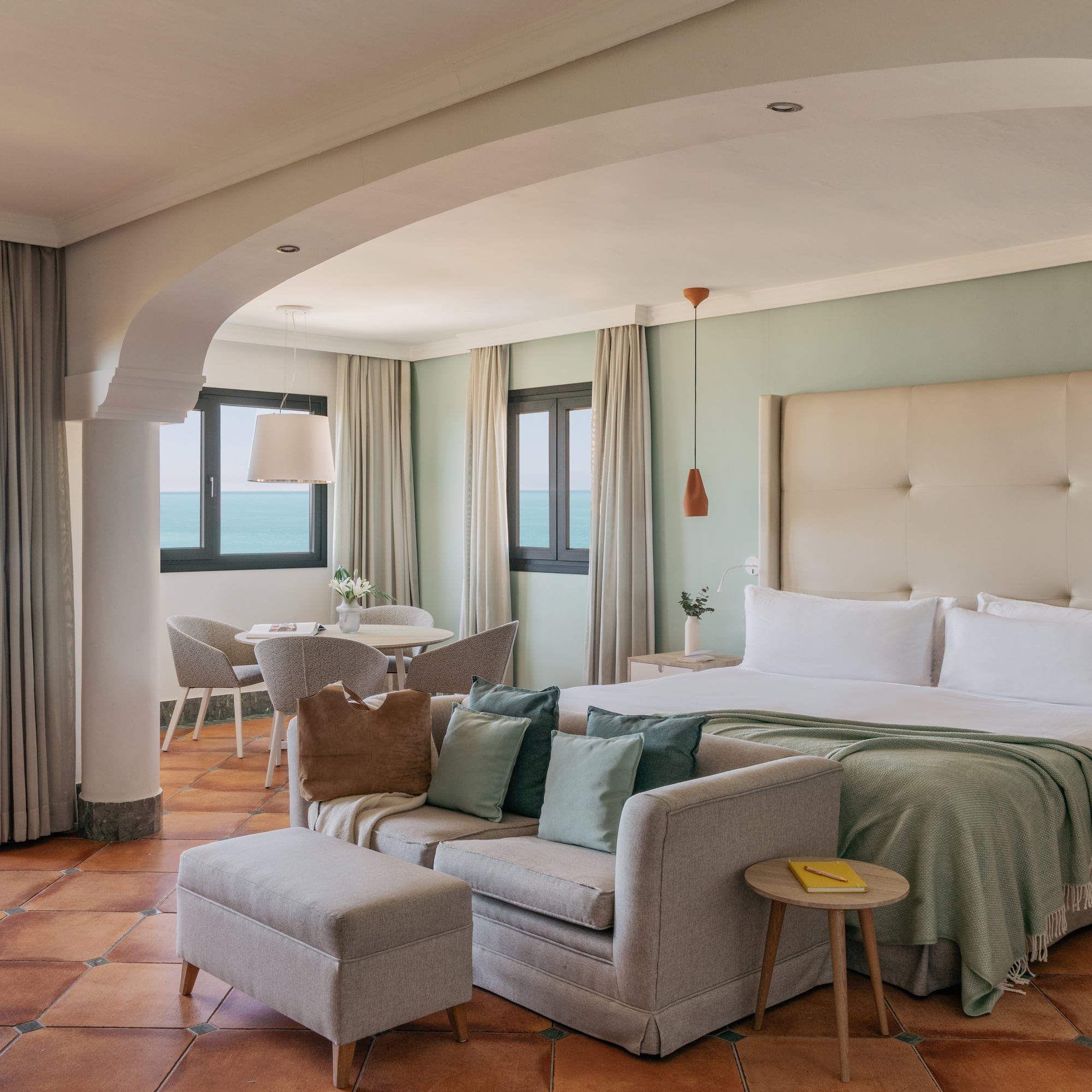 Coastal hotel suite with ocean view balcony, bed, sofa, and terracotta floor.