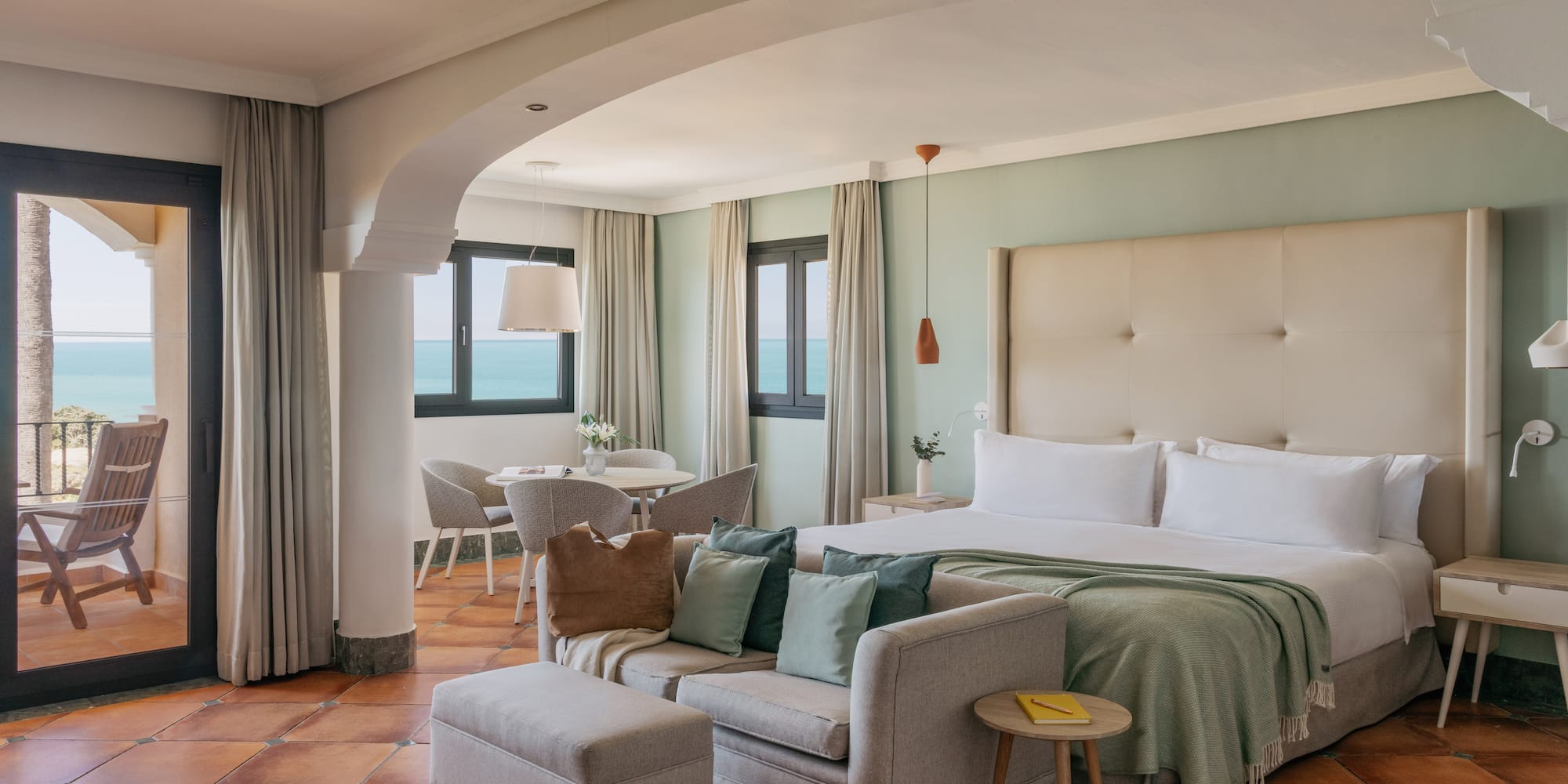 Coastal hotel suite with ocean view balcony, bed, sofa, and terracotta floor.