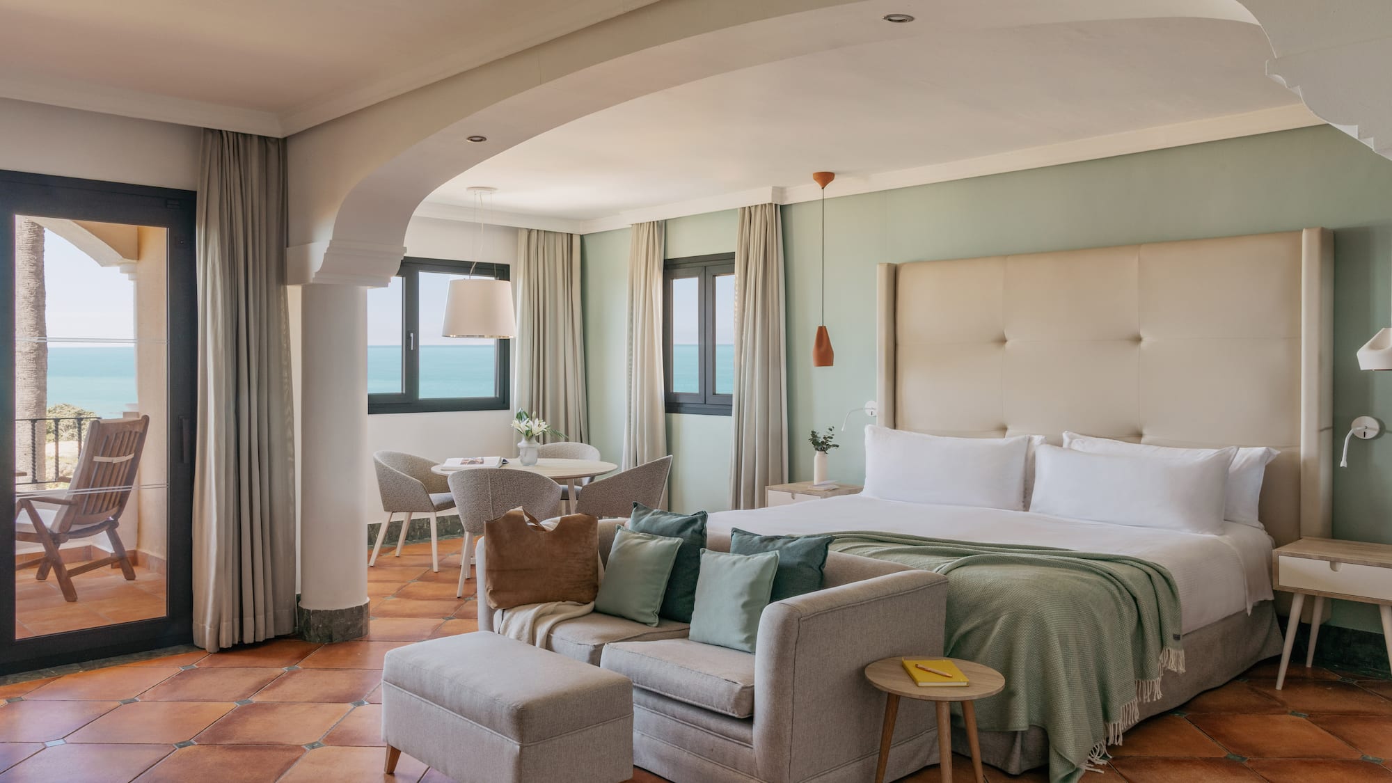 Coastal hotel suite with ocean view balcony, bed, sofa, and terracotta floor.