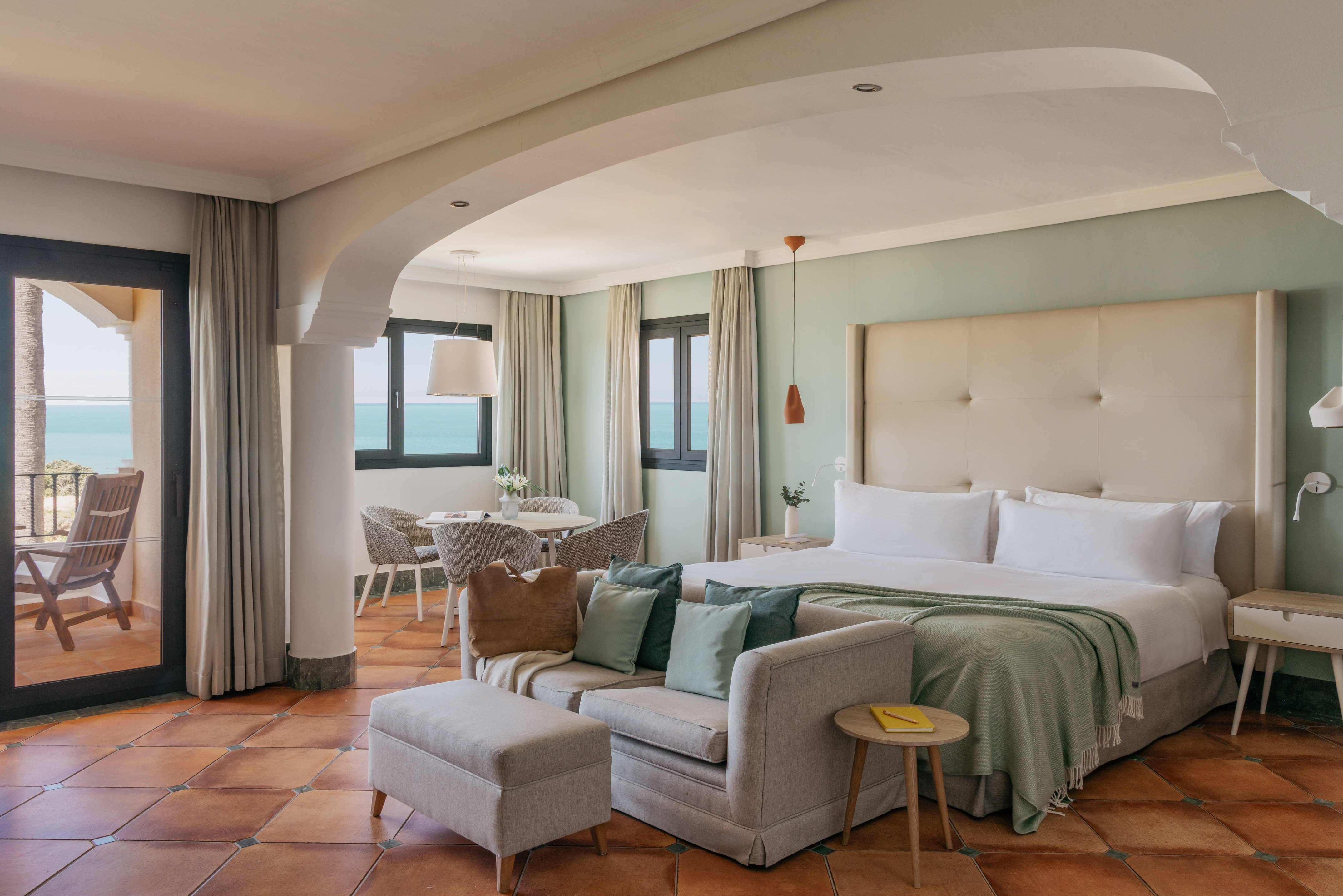 Coastal hotel suite with ocean view balcony, bed, sofa, and terracotta floor.