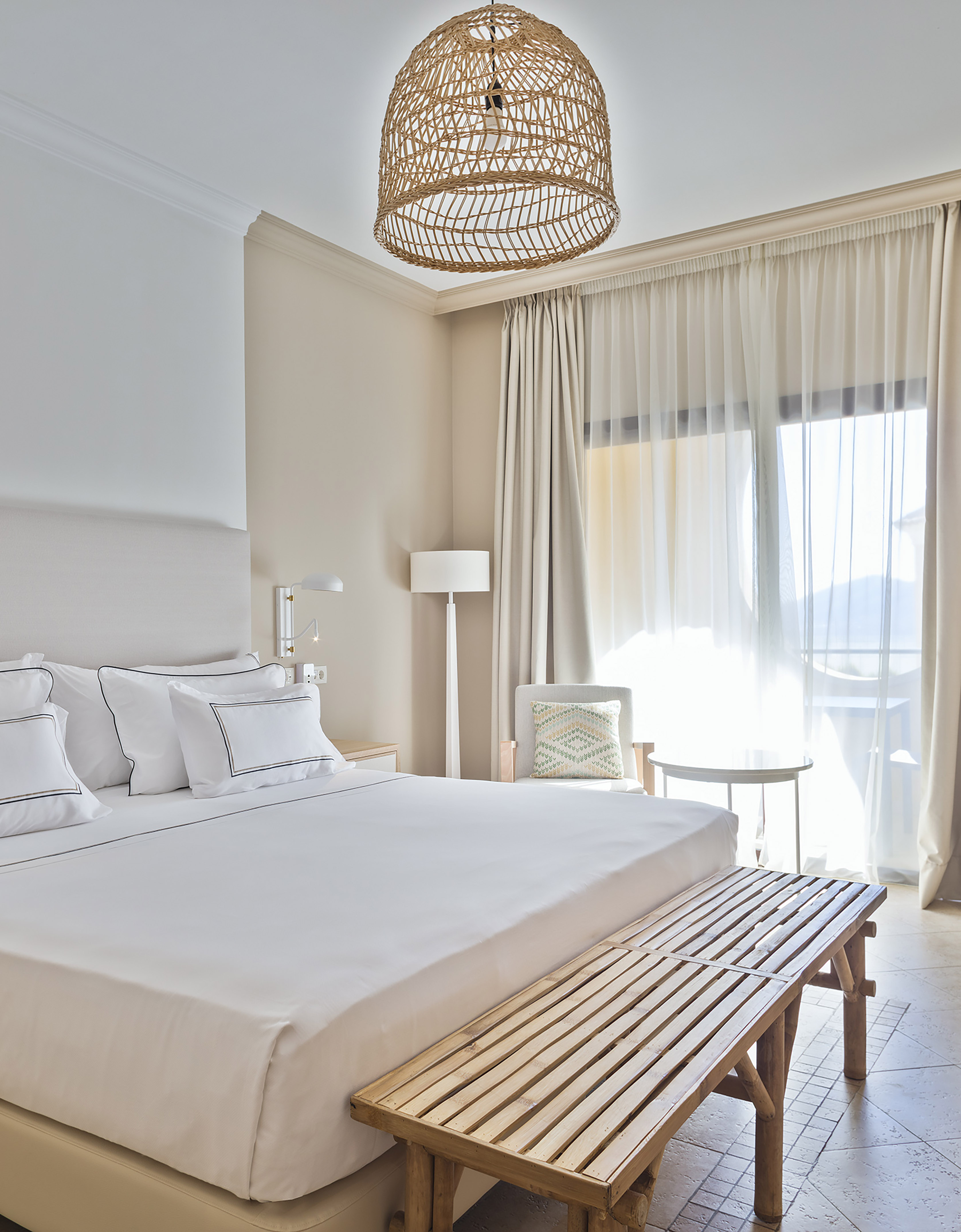 Modern bedroom with white bed, wicker pendant, wood bench, and sunlit window.
