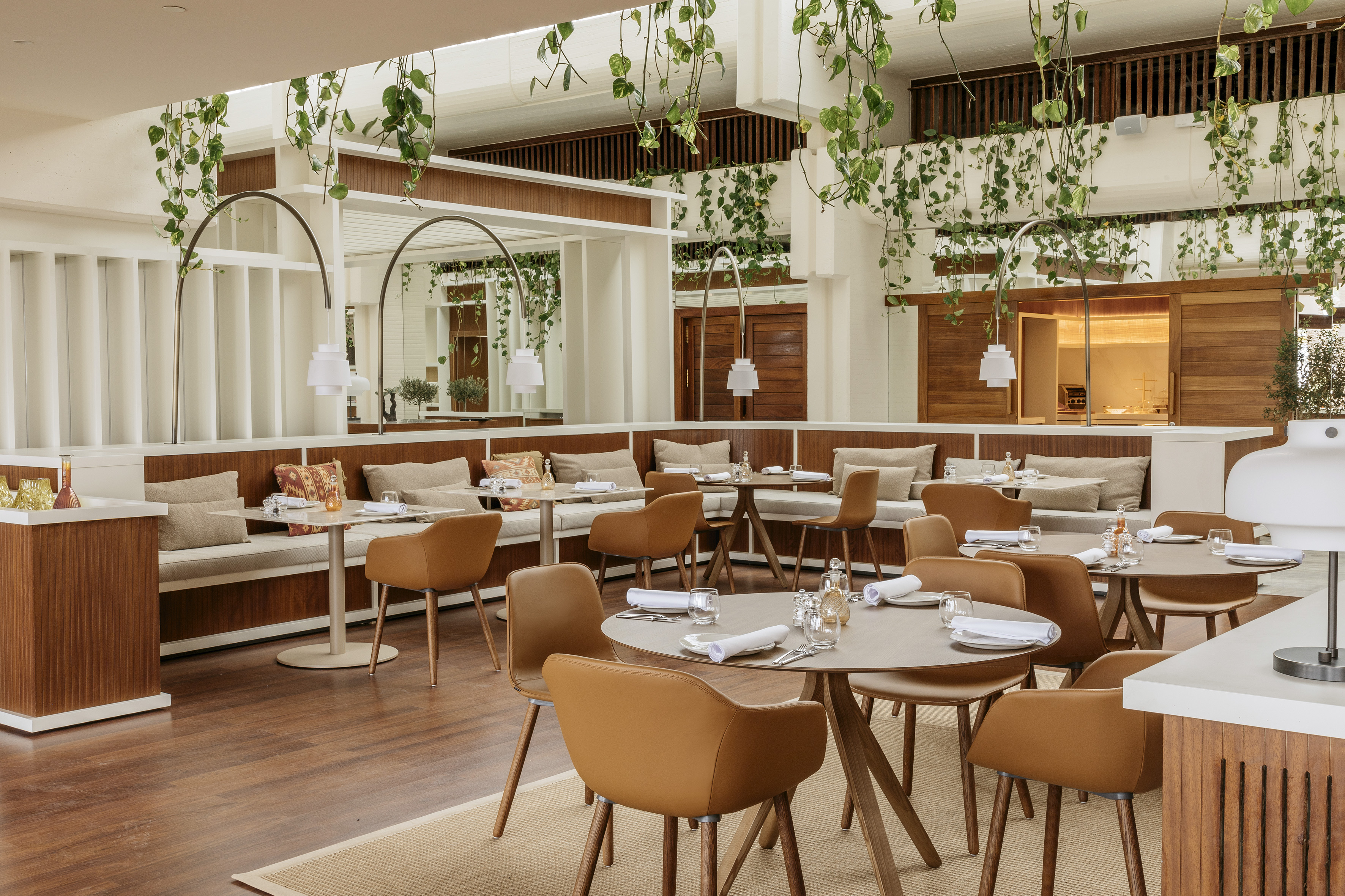 Modern restaurant dining area with wood, tan seating, and lush hanging plants.