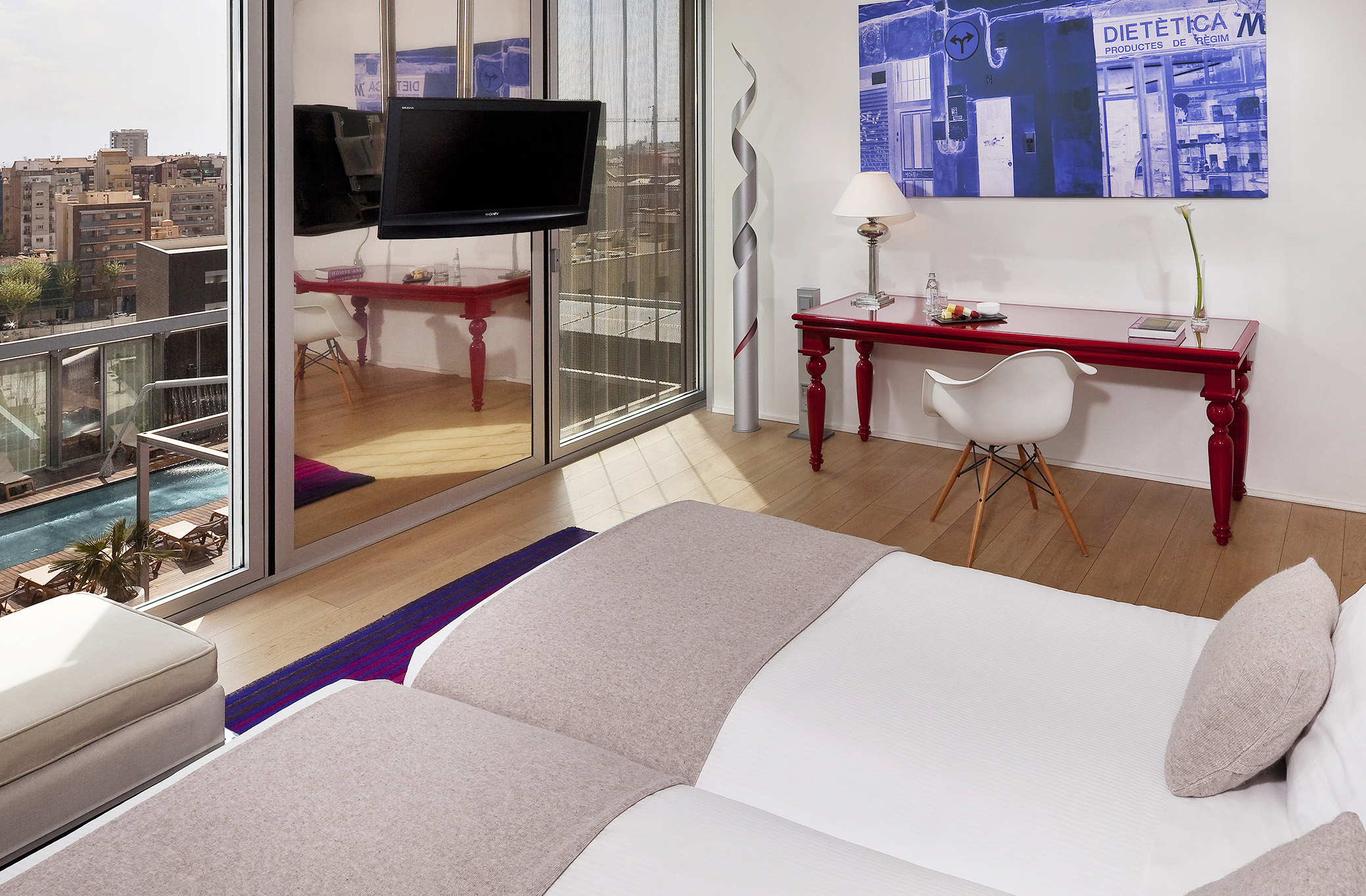Modern hotel room with twin beds, red desk, city view balcony, and outdoor pool.