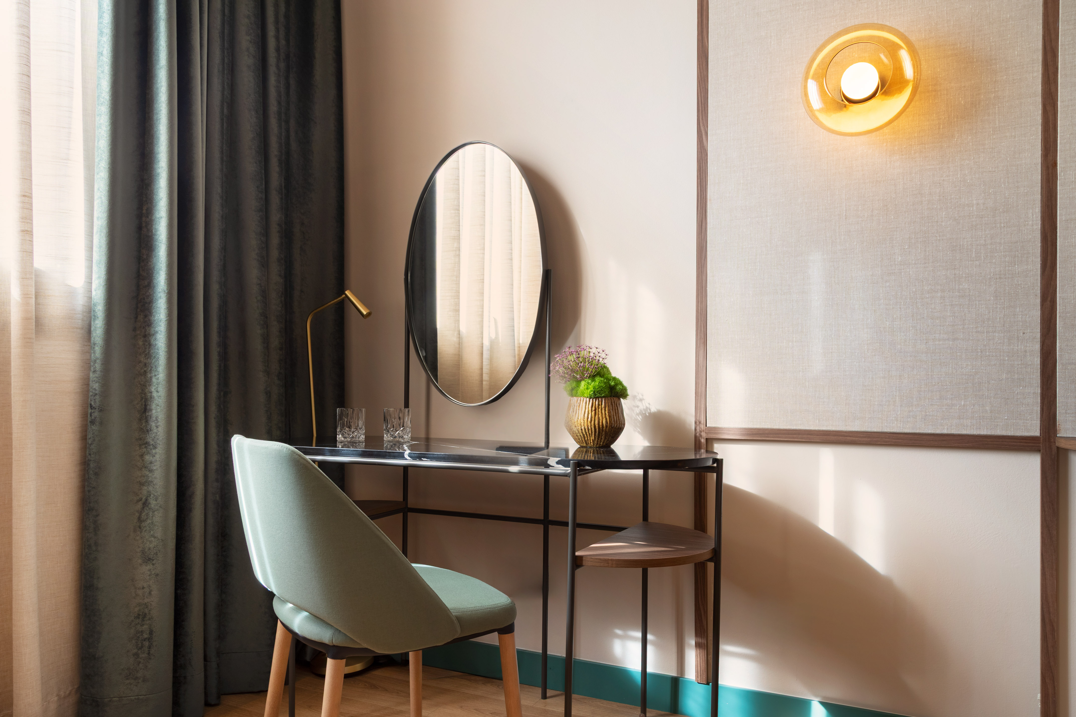 Modern hotel vanity with oval mirror, green chair, gold lamp, and wall light.