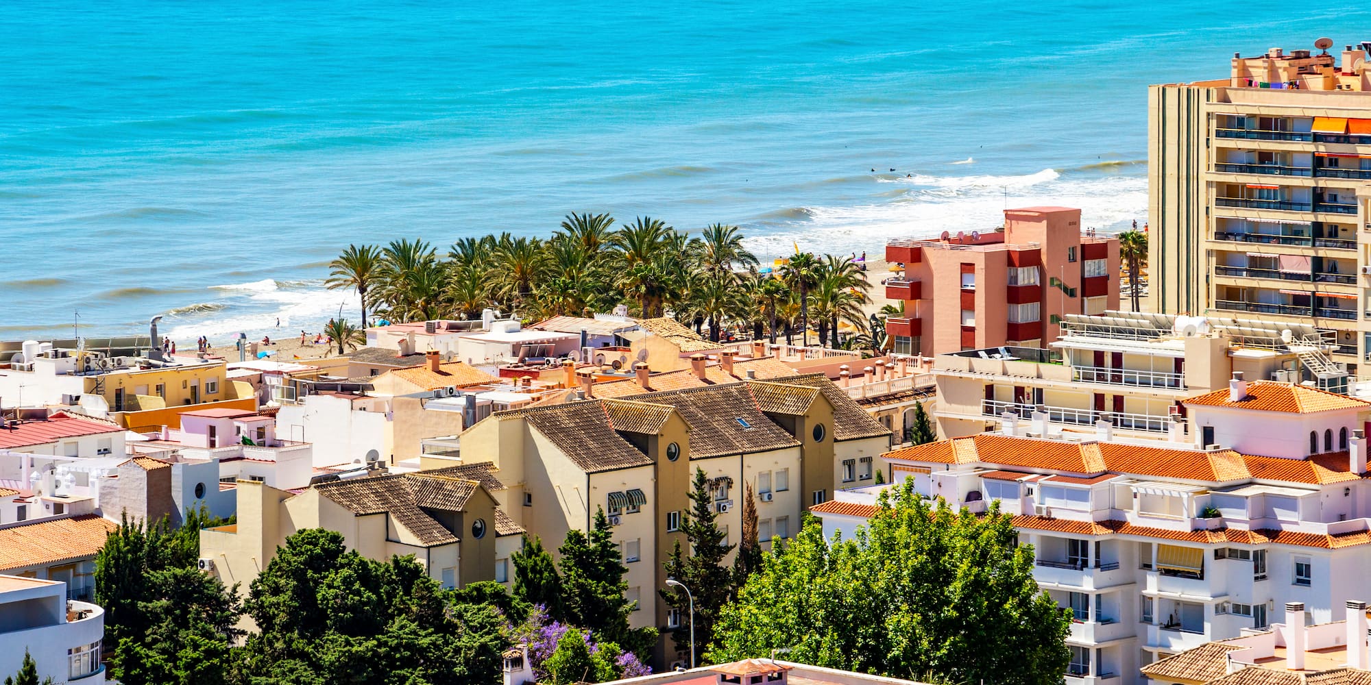 Sunny coastal town with turquoise sea, beach, palm trees, and diverse buildings.