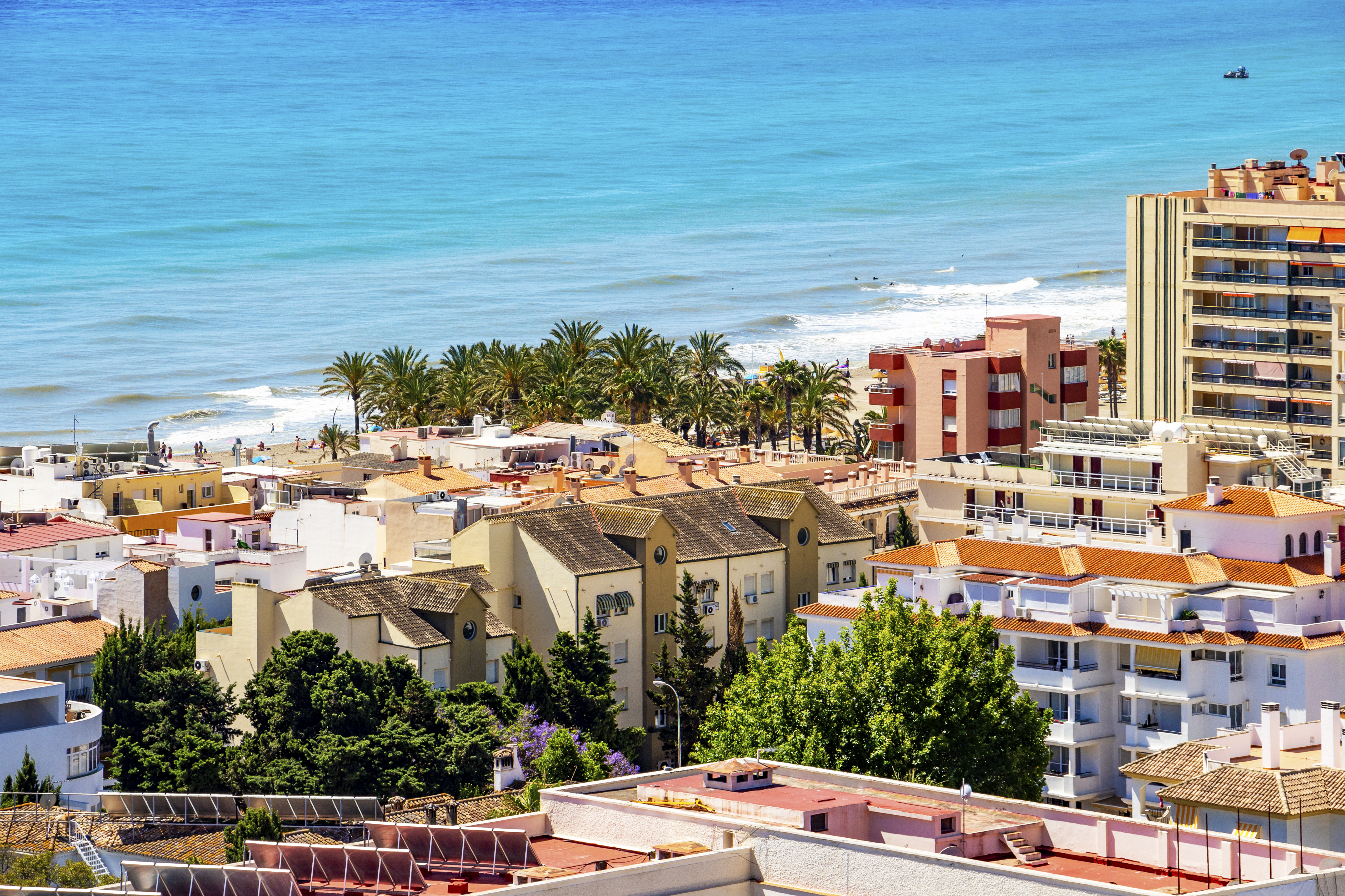 Sunny coastal town with turquoise sea, beach, palm trees, and diverse buildings.
