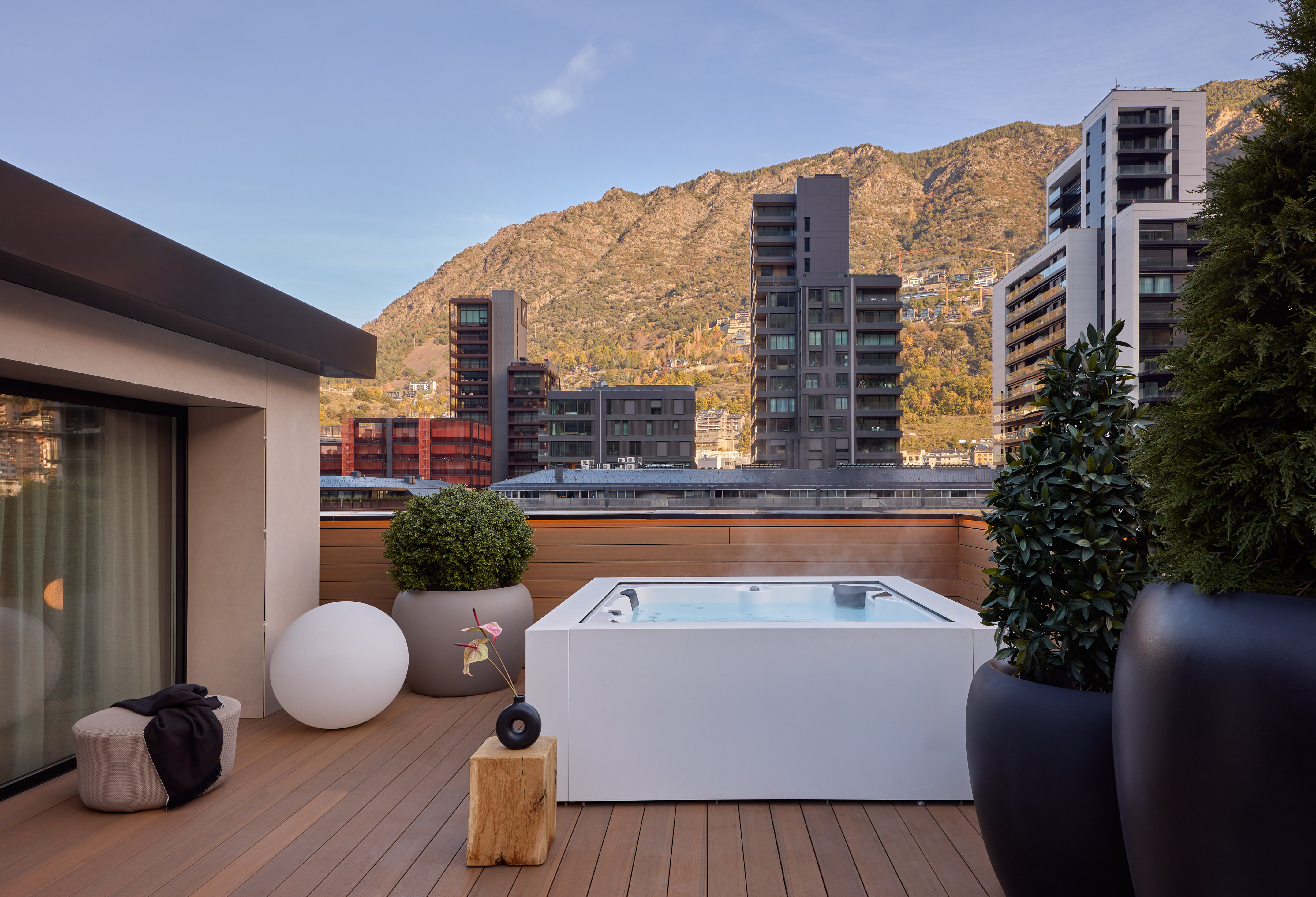 Modern rooftop terrace with hot tub, city buildings, and mountain backdrop.