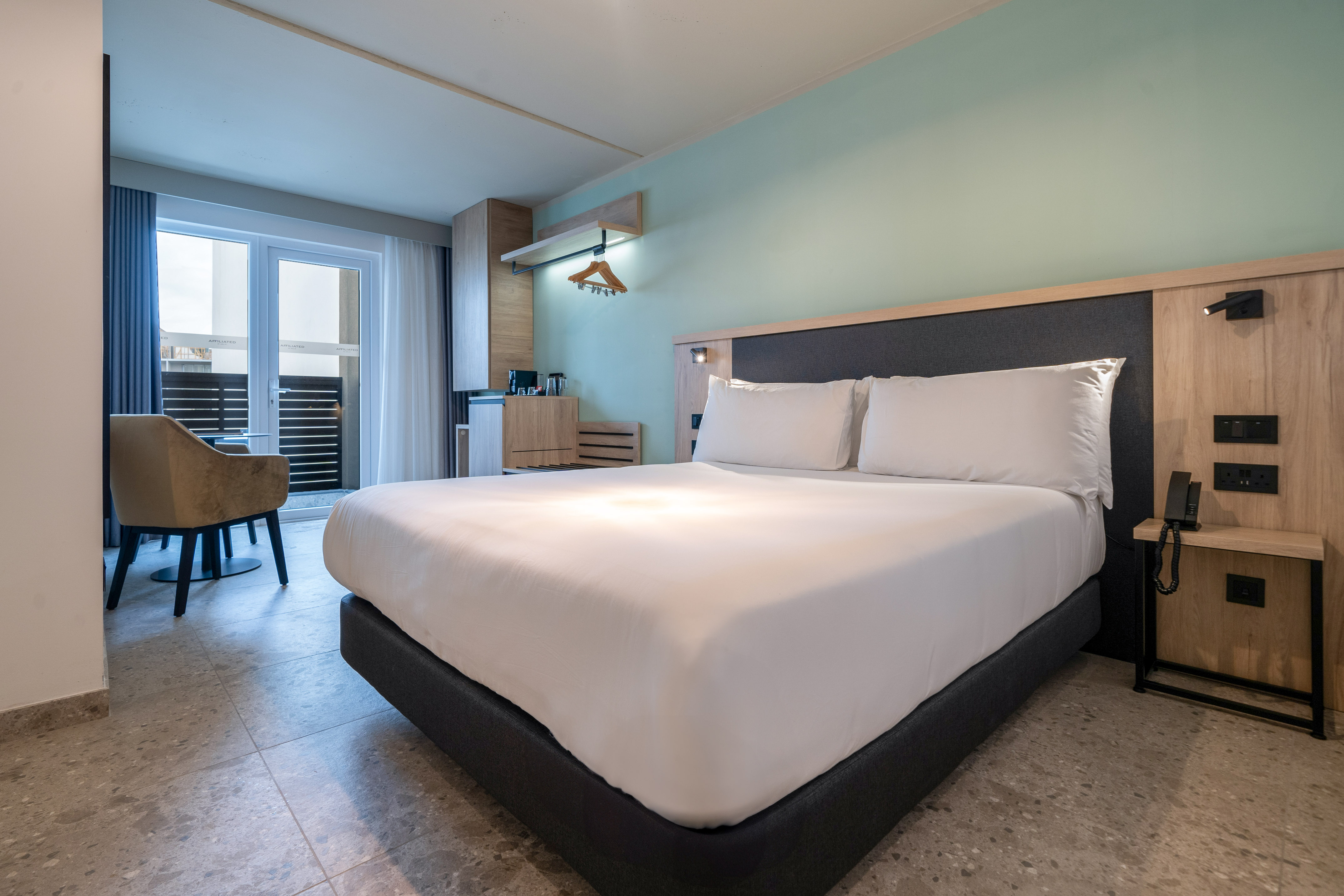 Modern hotel room with king bed, balcony access, and light green walls.