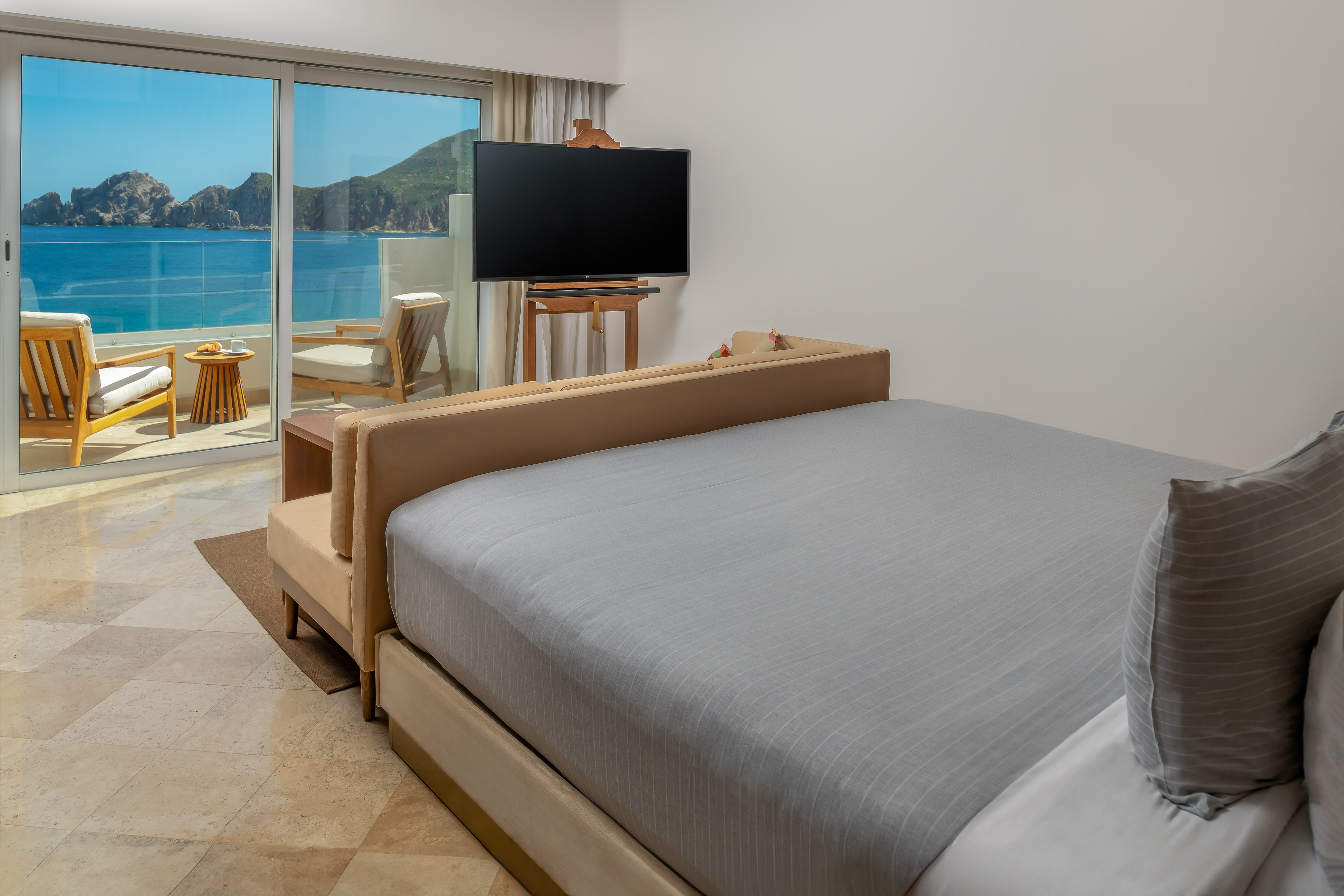 Luxury hotel room with king bed, ocean view balcony, TV, and distant cliffs.
