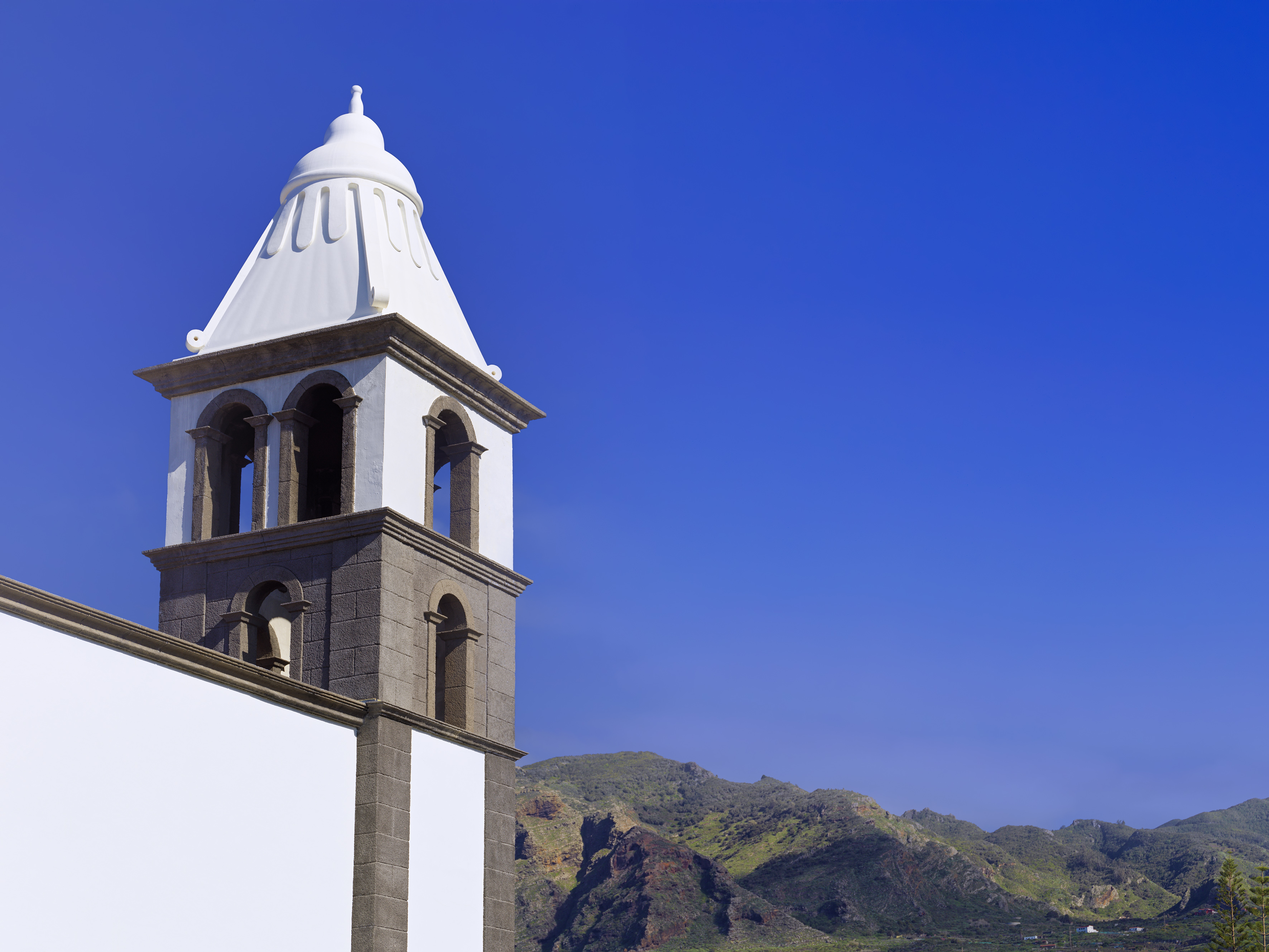 White church bell tower with stone base, green mountains, and blue sky.