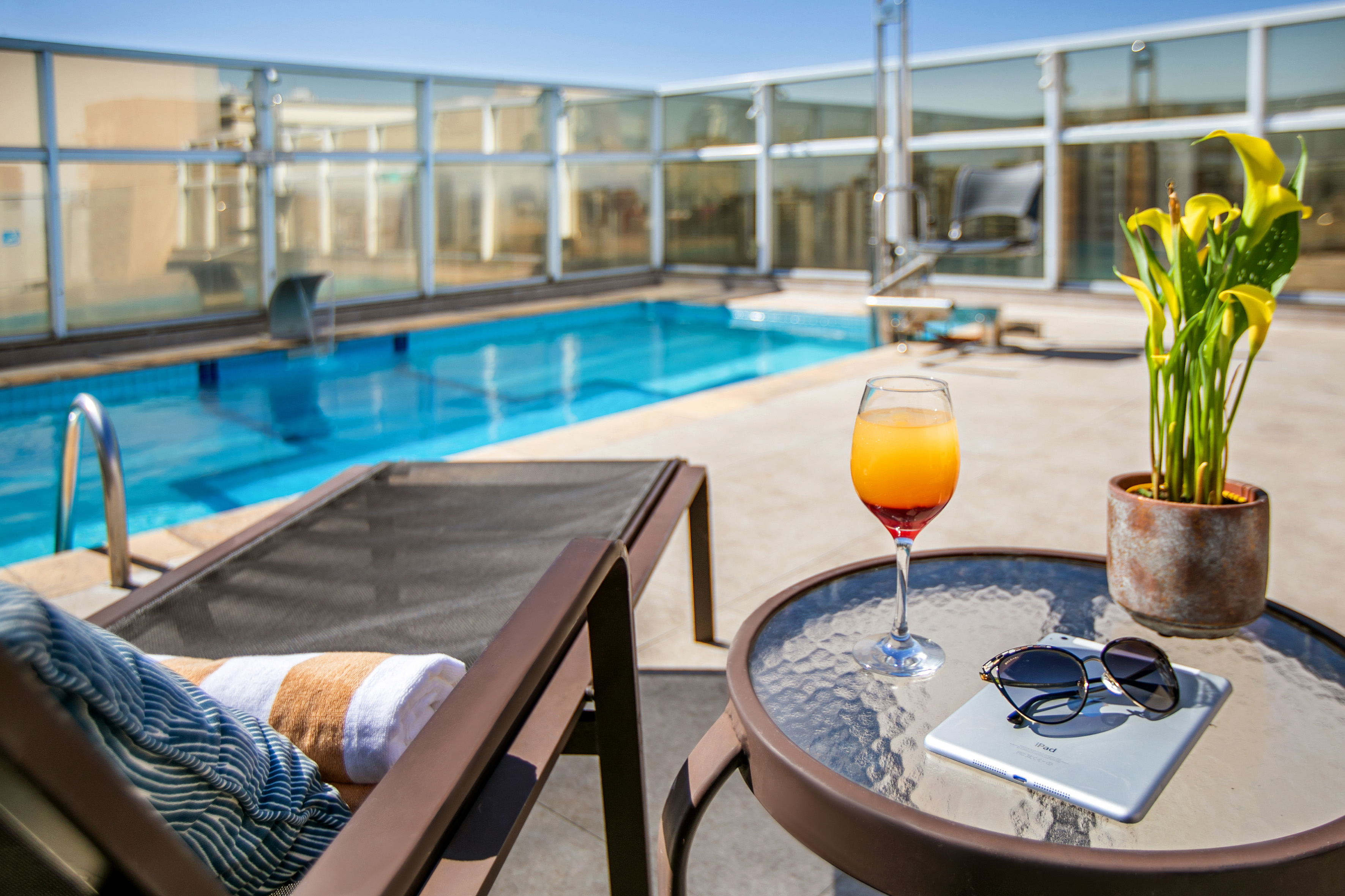 Rooftop hotel pool, lounge chair, cocktail, iPad, and sunglasses on a sunny day.